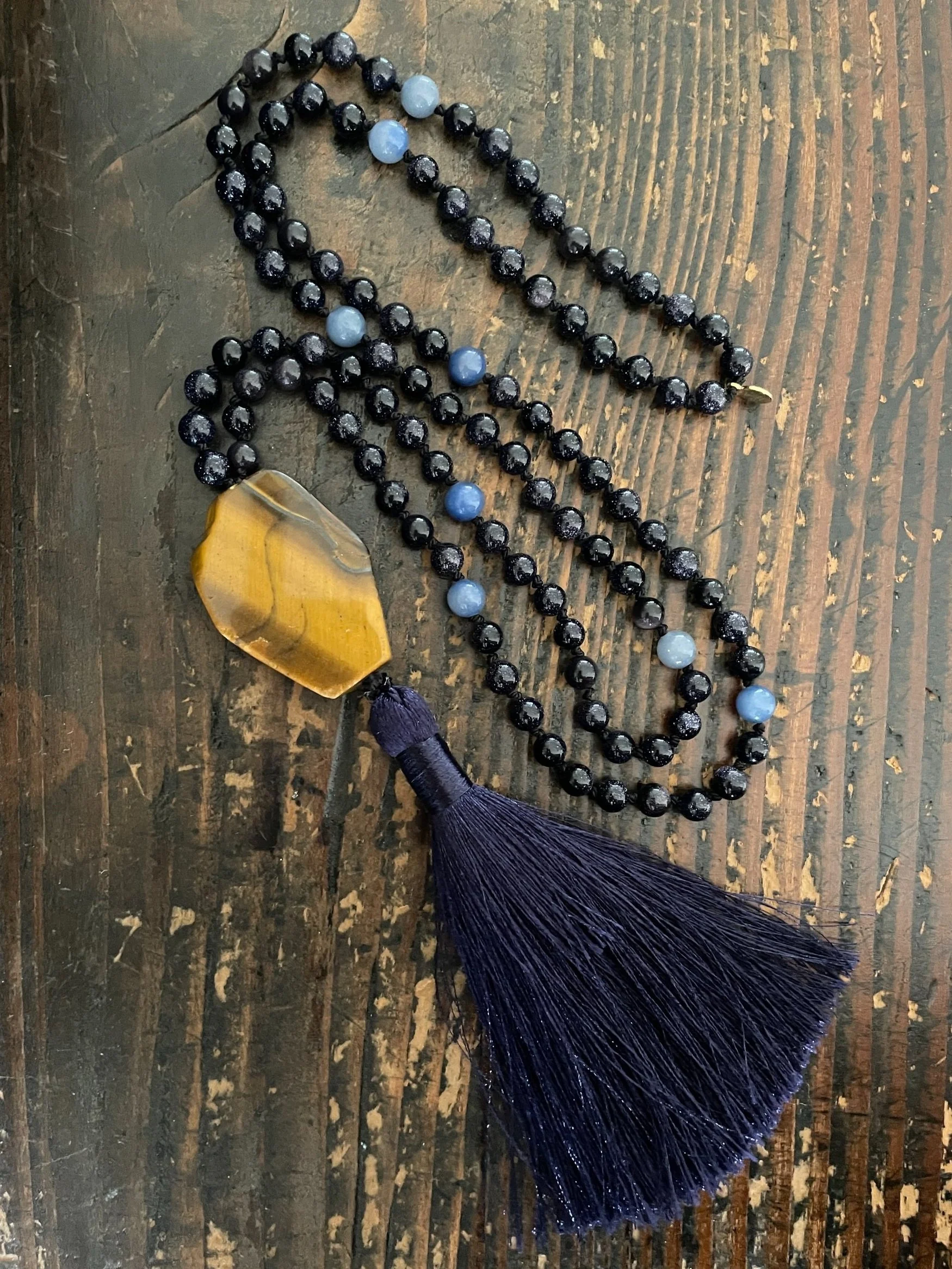 A beaded necklace with a large yellow/golden pendant and a dark blue tassel. The necklace includes black beads and light blue beads, lying on a wooden surface.