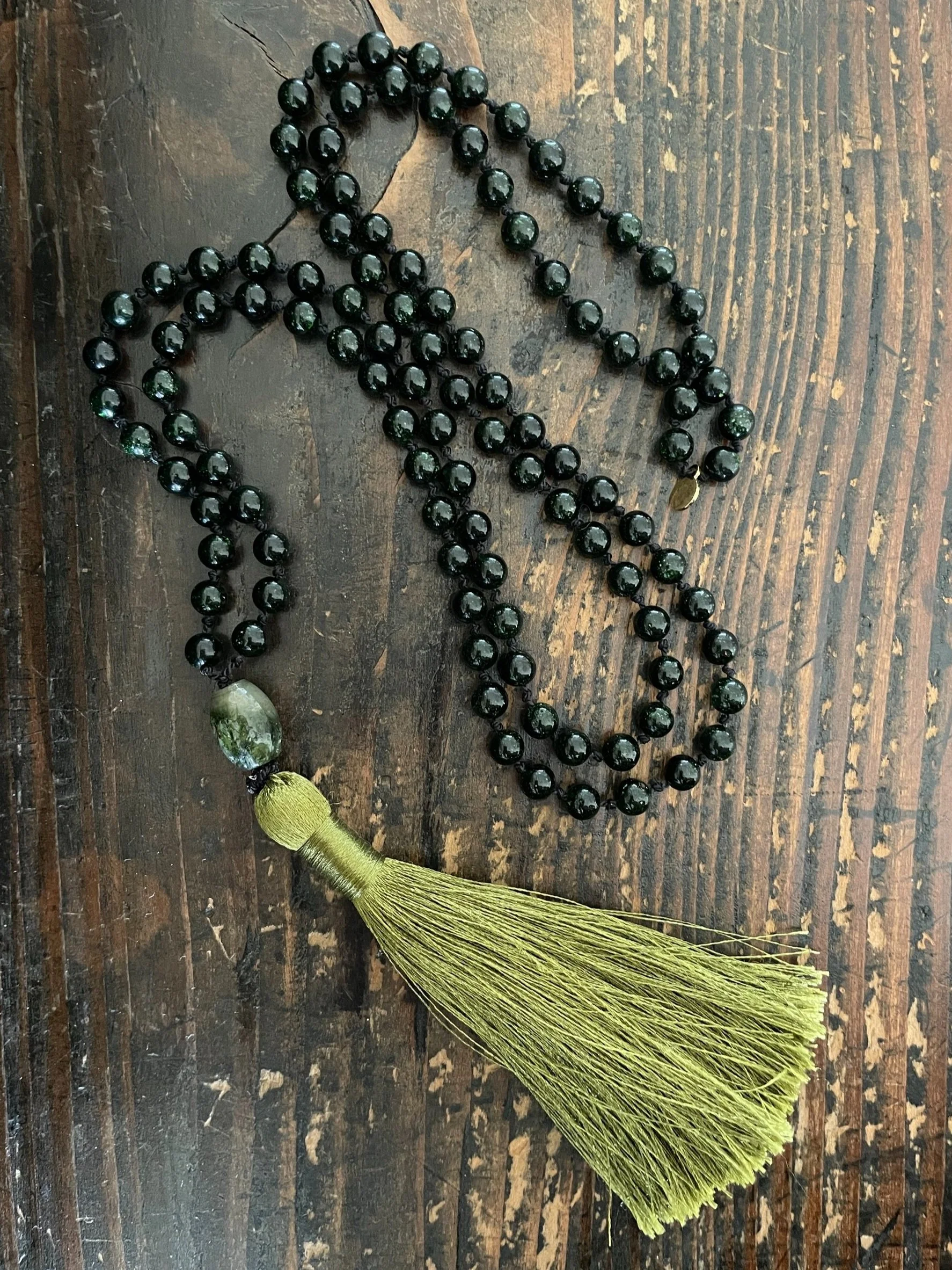 Green goldstone mala necklace with a moss agate bead and a gold tassel, placed on a wooden surface.