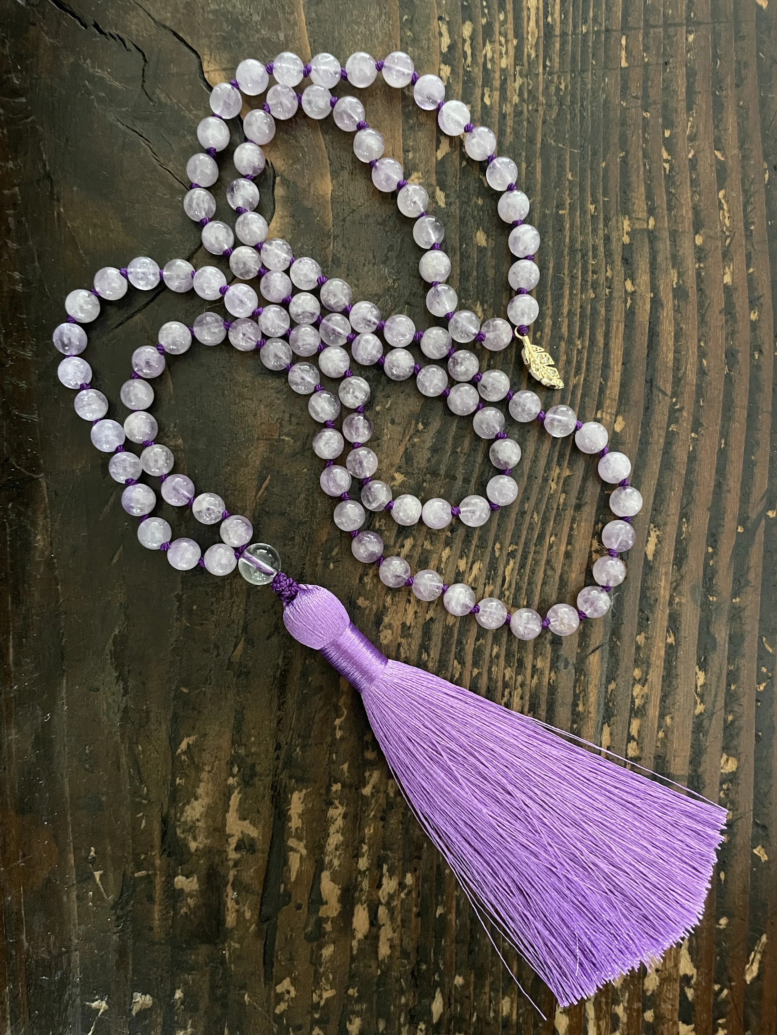 A lilac amethyst beaded mala with a purple tassel resting on a dark wooden surface.