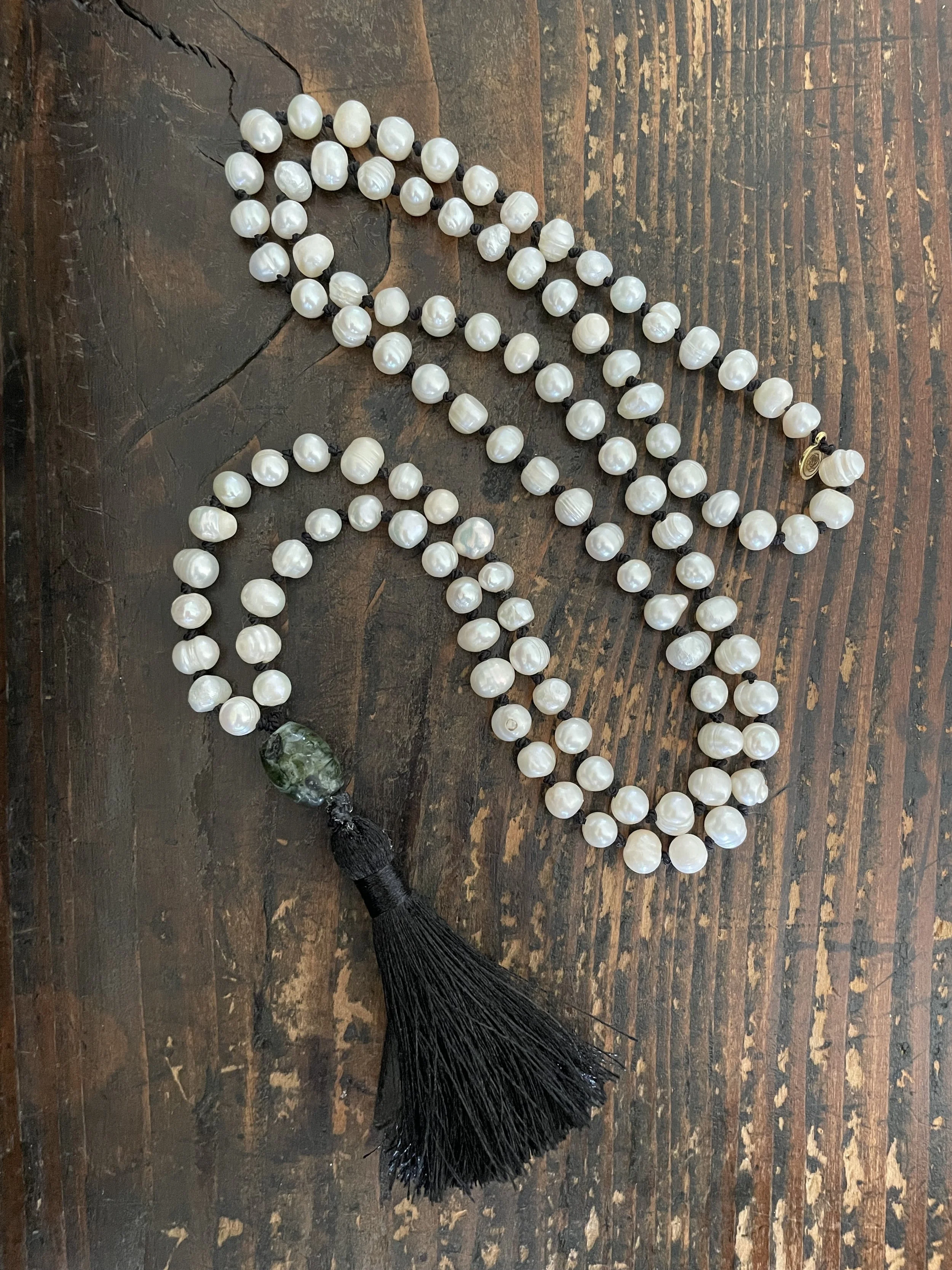 A string of white pearl beads with a black tassel and a green moss agate bead, laid on a wooden surface.