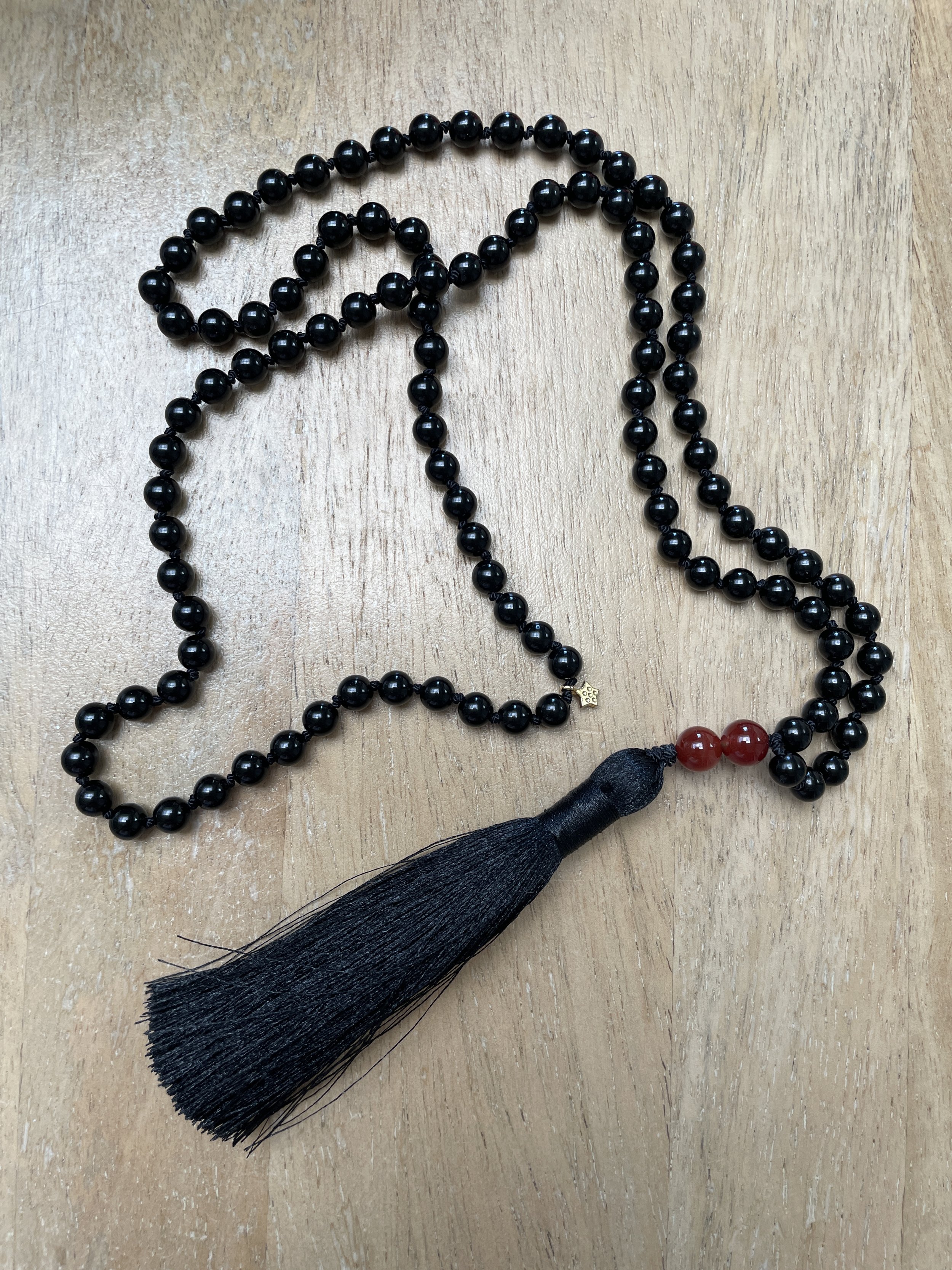 A black beaded meditation mala with a black tassel and two red beads near the tassel, lying on a wooden surface.