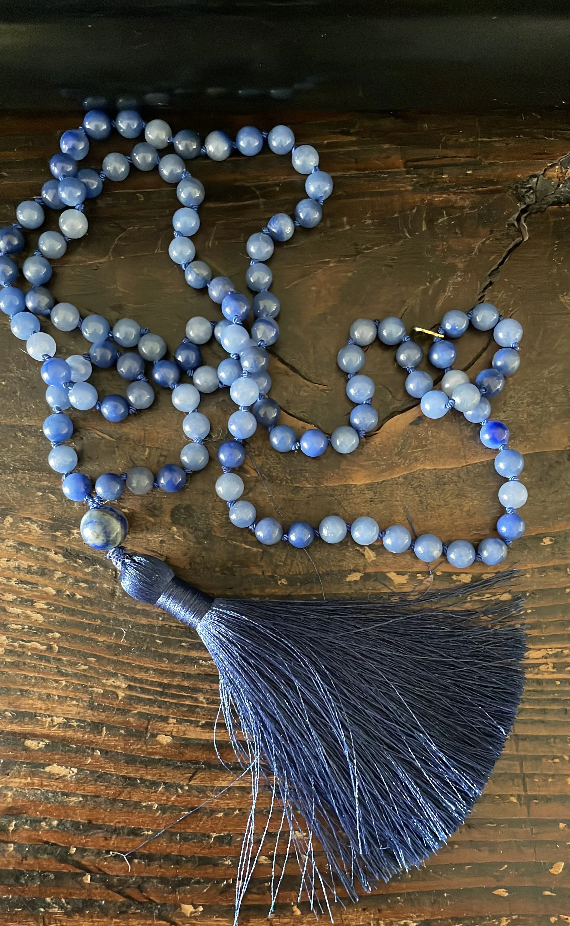 A necklace made of blue aventurine beads with a lapis lazuli bead, dark blue tassel lying on a wooden surface.