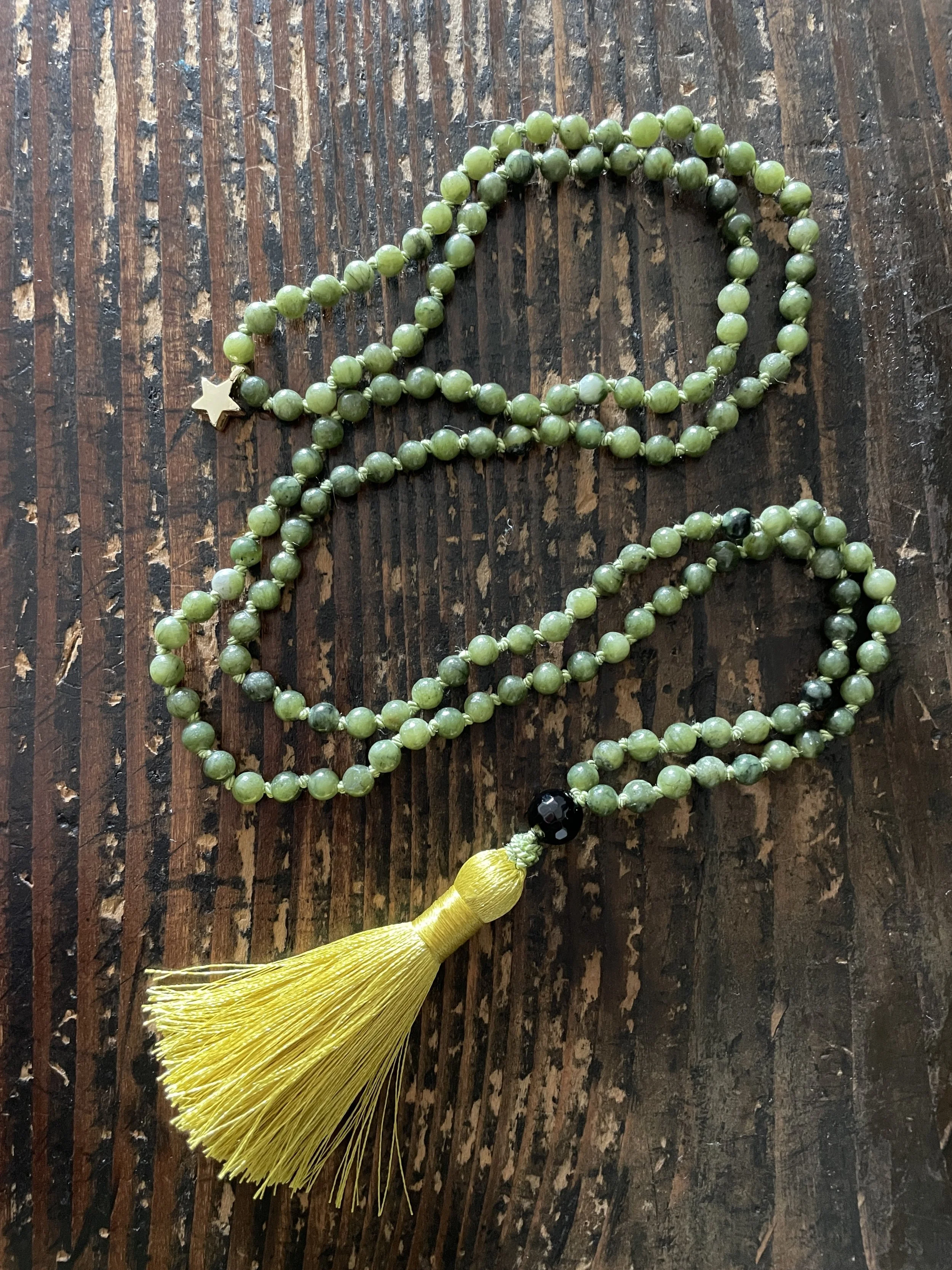 Green nephrite jade beads with a black onyx bead, yellow tassel and a small gold plated star charm on a wooden surface.