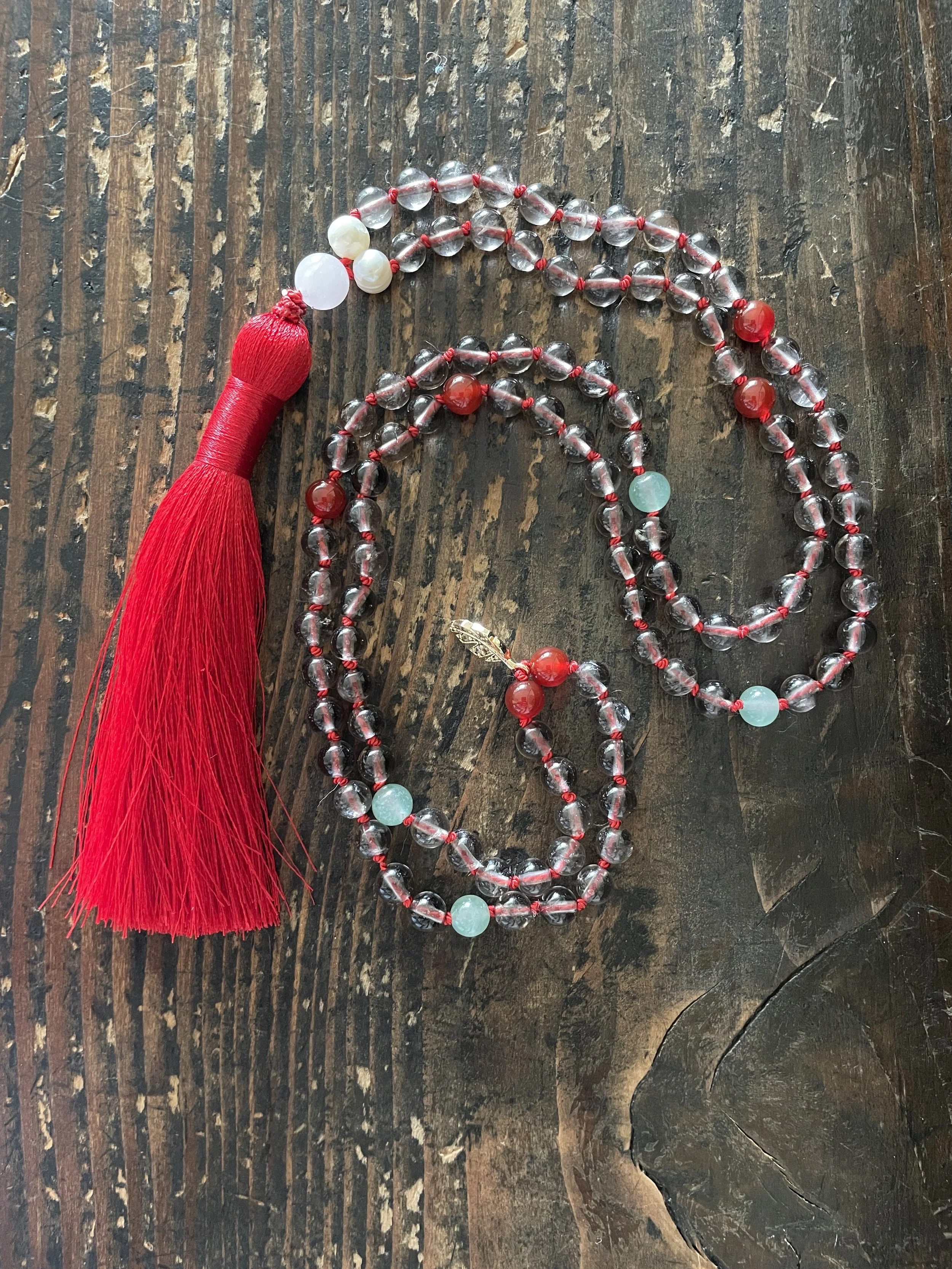 A beaded meditation necklace with a red tassel, featuring clear and red beads, and some light green beads on a wooden surface.