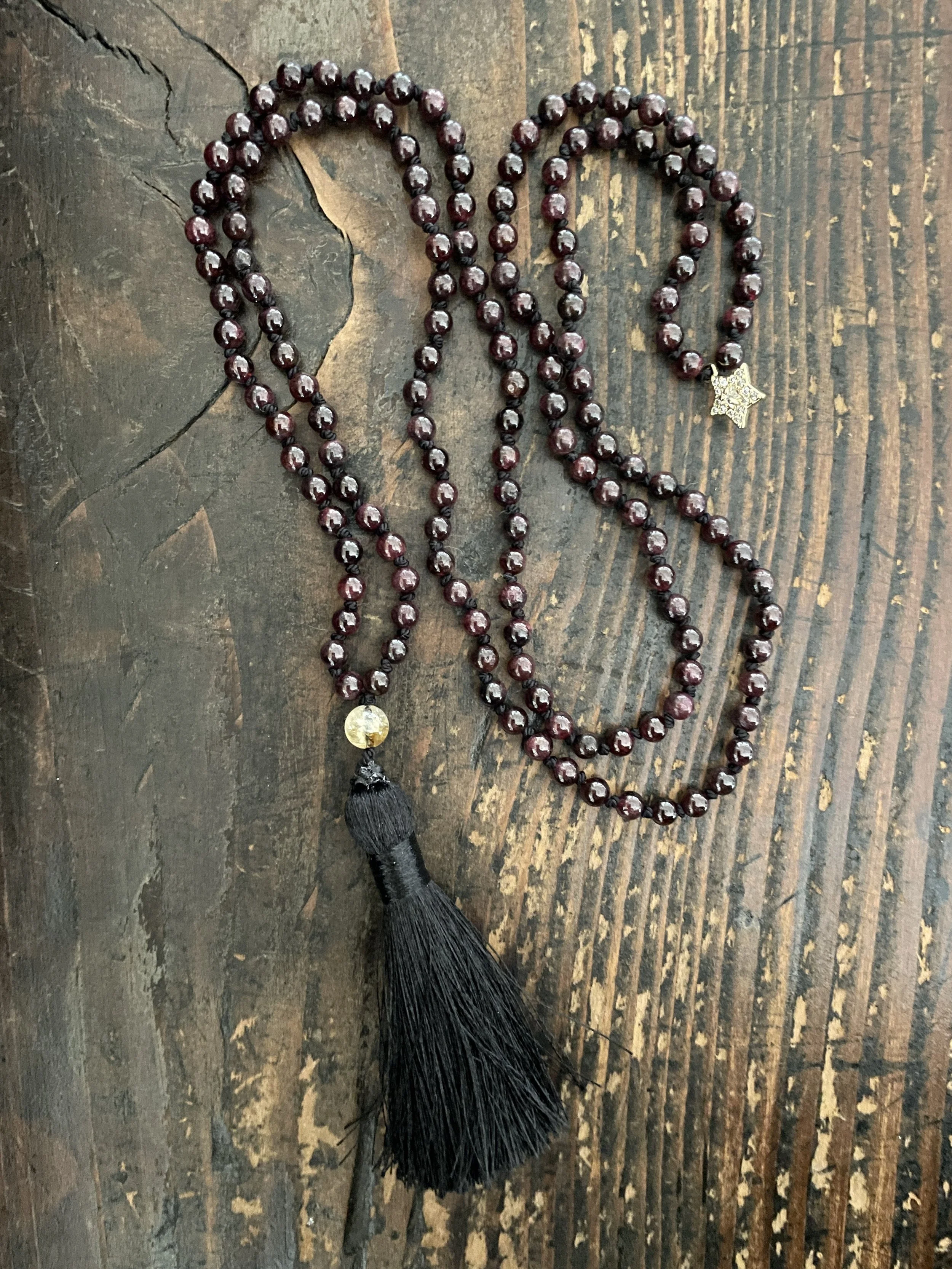 A mini meditation mala with 152 garnet beads, featuring a black tassel and a small gold star-shaped charm, laid on a wooden surface.