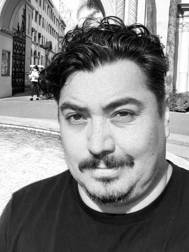 Close-up of a man with dark, curly hair and facial hair, smiling slightly, outdoors near a blue swimming pool with a cityscape background.