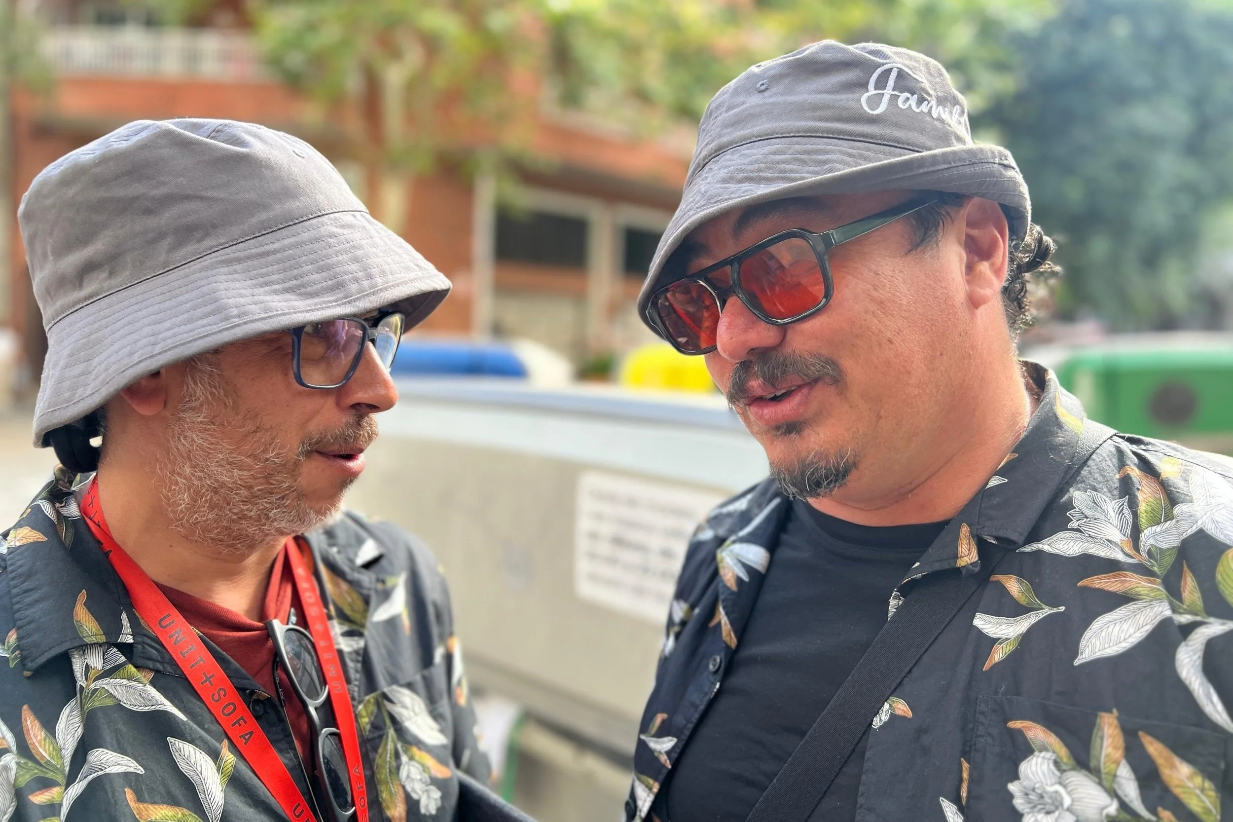 Two men wearing grey bucket hats, Hawaiian shirts, and sunglasses are engaged in conversation outdoors.
