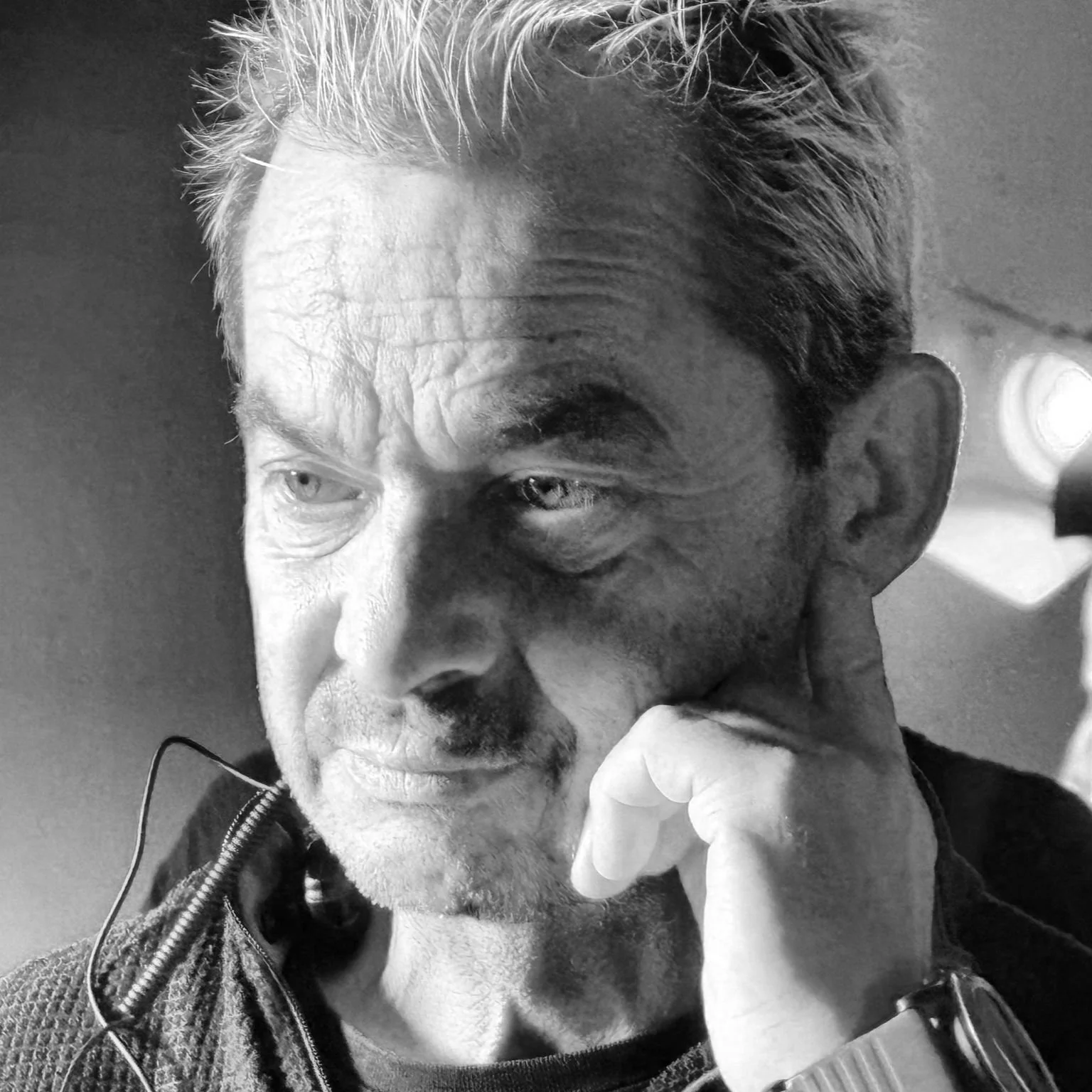 Close-up of an older man with grayish hair, wearing a dark jacket and a wristwatch, resting his chin on his hand, with a thoughtful expression, colorful background, and a blue-green light on the right side.