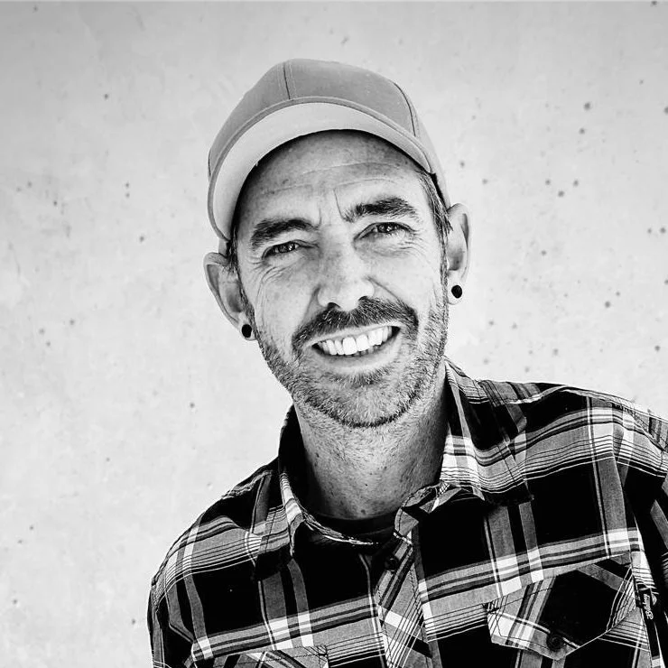 Black and white photo of a man wearing a baseball cap, plaid shirt, and smiling at the camera against a plain background.