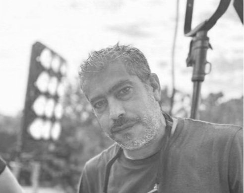 A man with short curly hair and stubble, wearing a dark T-shirt, standing outdoors with a lighting setup in the background.