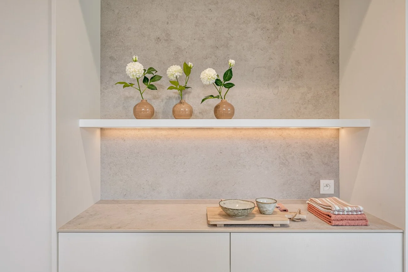A kitchen or dining area with three small vases holding white flowers on a white shelf, modern beige countertop with bowls and kitchen towels, and a textured beige wall with a power outlet, under soft lighting.
