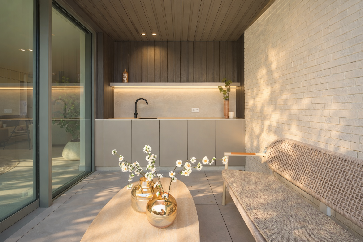 Sunlit outdoor patio area with a beige bench, a wooden table with white daisies in golden vases, a modern kitchenette with a black faucet, and a large glass sliding door reflecting part of the patio.