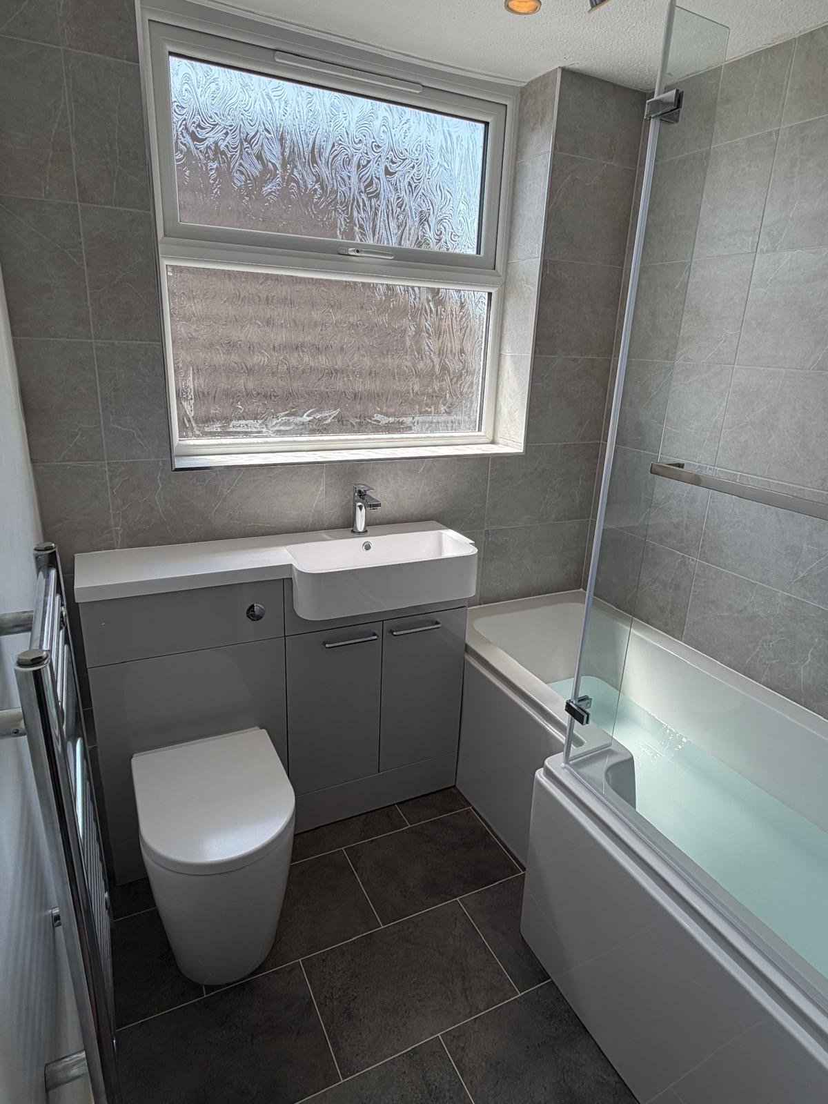 Small bathroom with a shower enclosure, small white sink with cabinet, a mirror, a frosted window, and a foldable shower seat inside the shower.