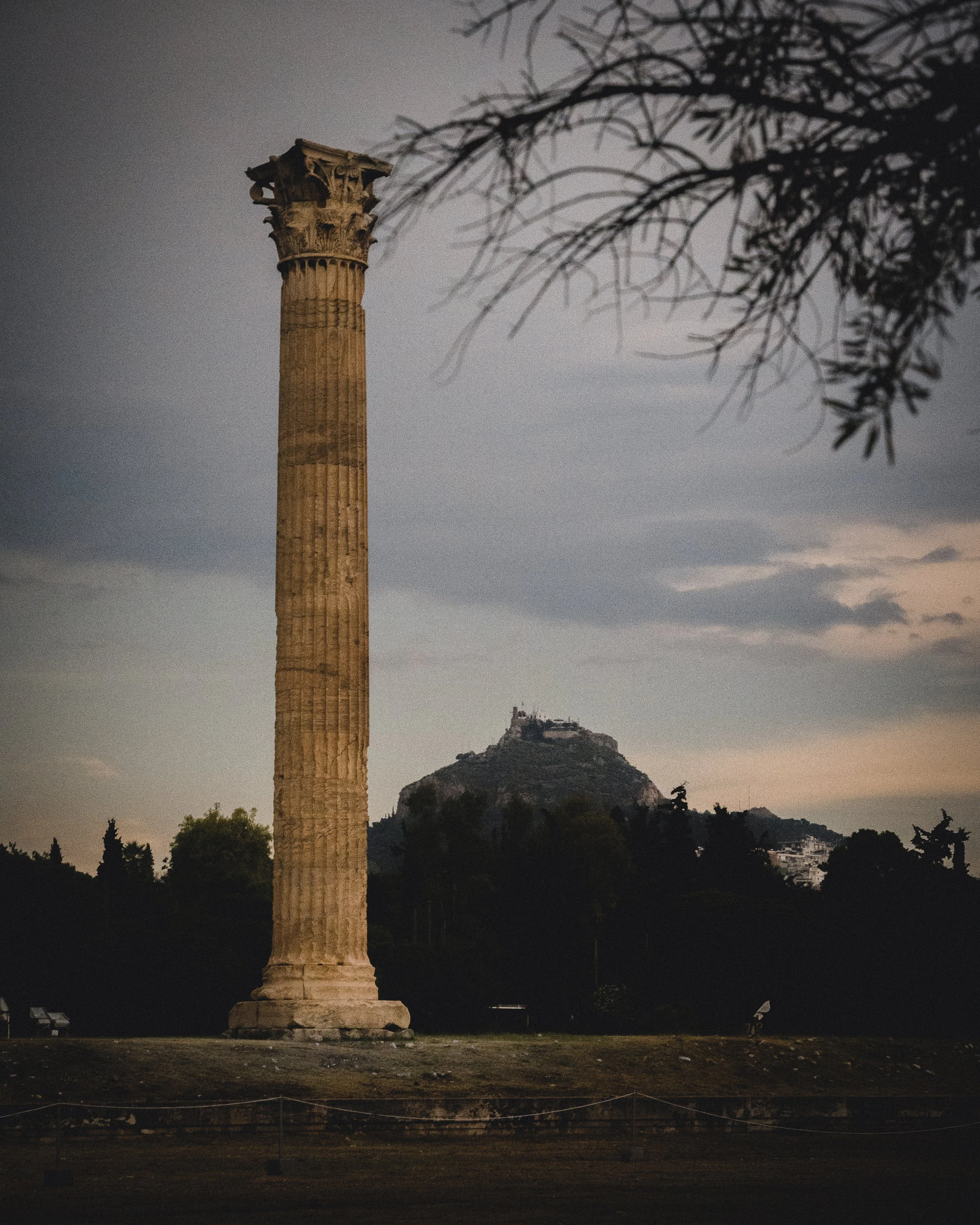 Colonne antique devant une colline au crépuscule, ciel nuageux et des arbres en arrière-plan.