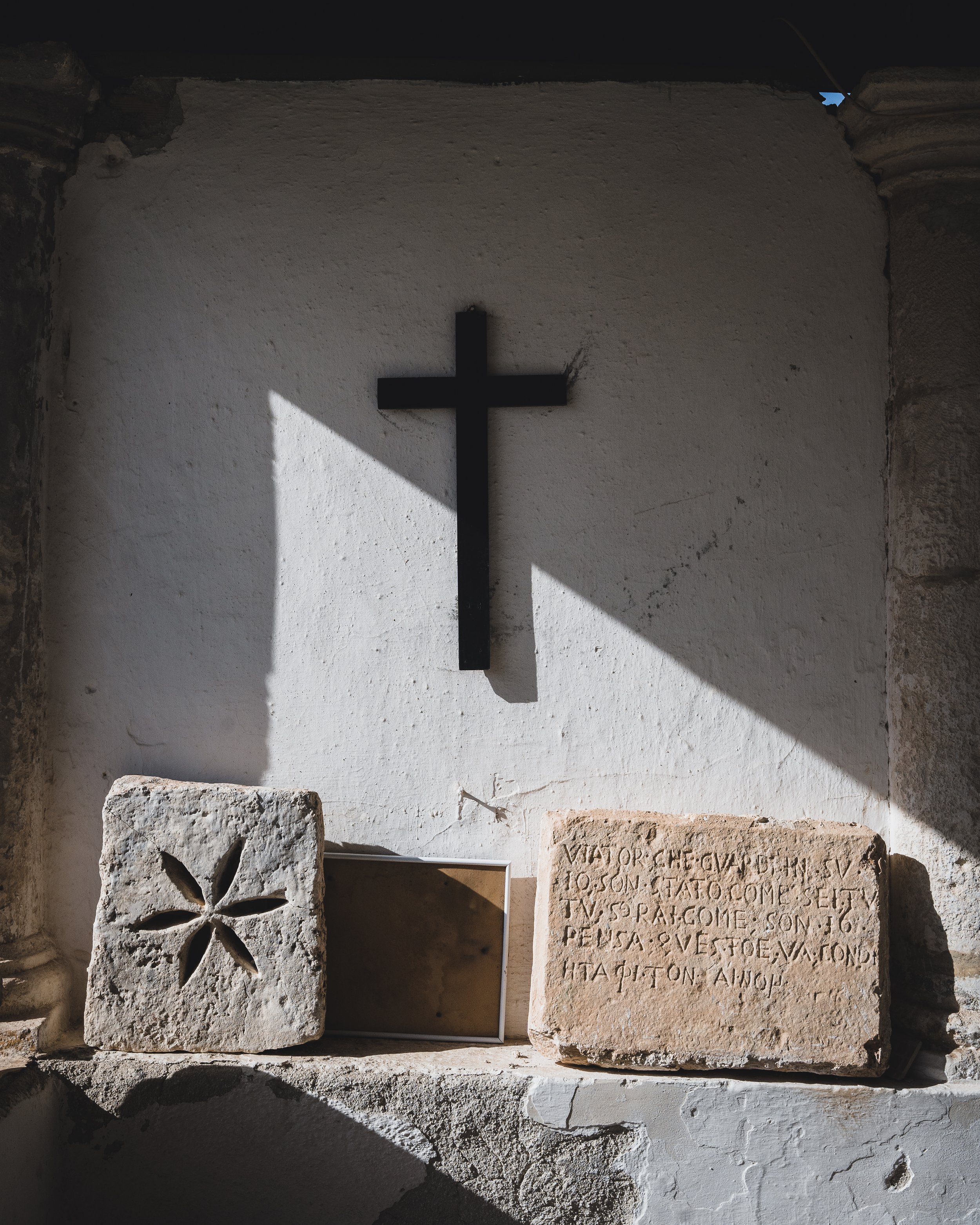 Une croix en métal est accrochée à un mur blanc. En dessous, il y a deux pierres anciennes avec des inscriptions et des motifs sculptés, posées sur une étagère en pierre éclairée par des rayons de soleil.