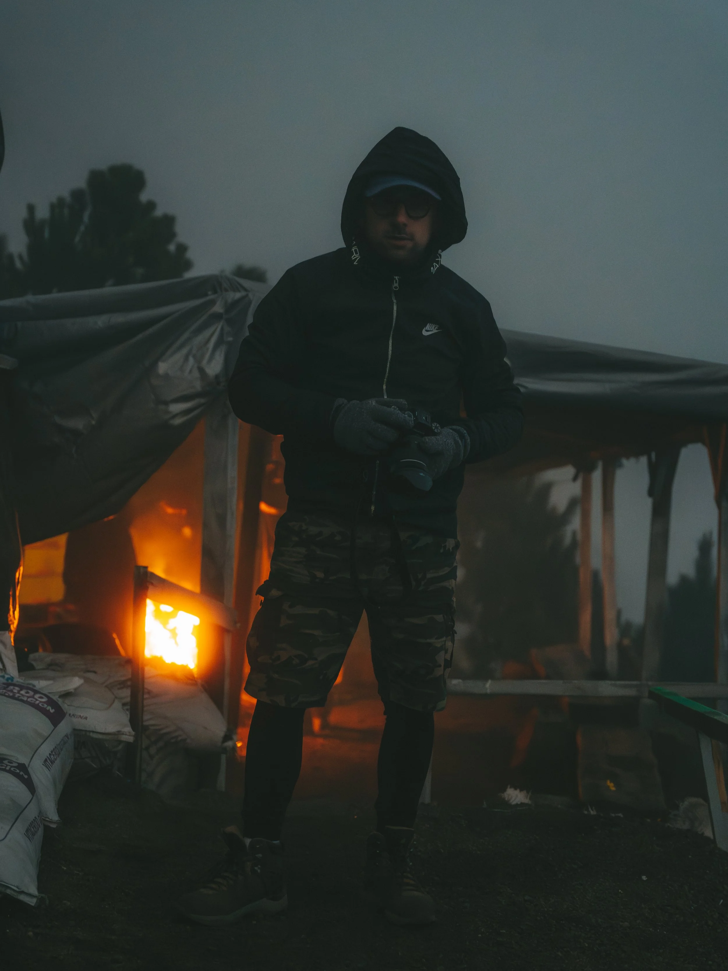 Un homme portant un manteau et un short camouflé se tient dehors, tenant un appareil photo devant une structure avec un feu à l'arrière-plan.