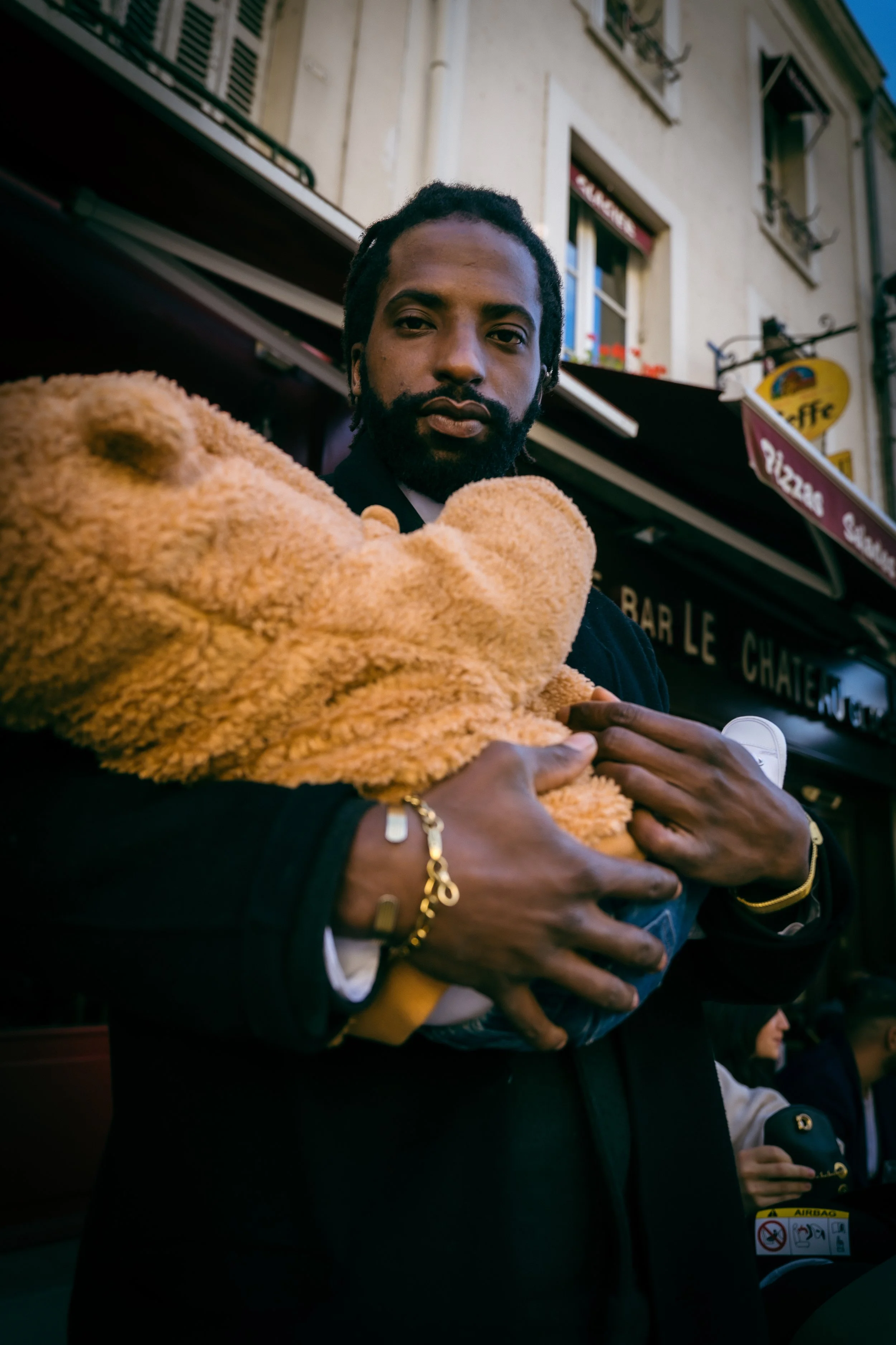 Un homme portant un gros ours en peluche dans une rue avec des bâtiments en arrière-plan.