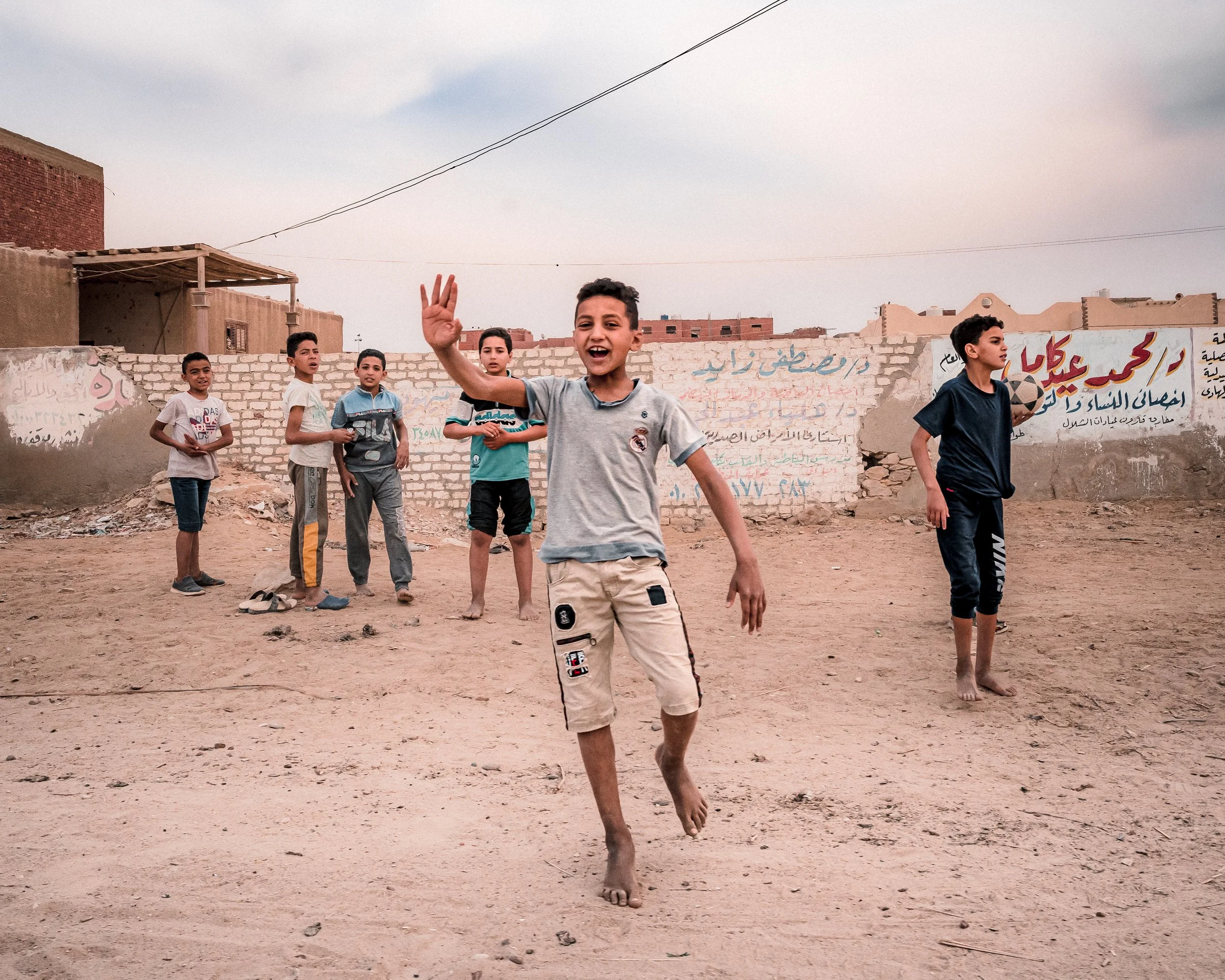 Un groupe d'enfants joue pieds nus dans un terrain poussiéreux devant un mur avec des graffitis en arabe.