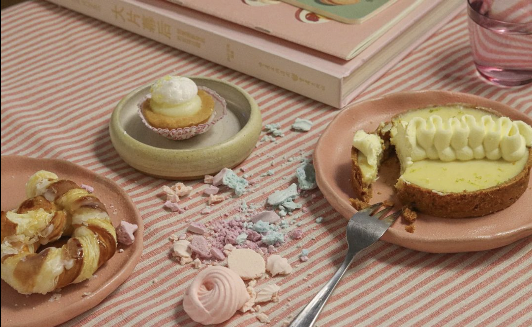 Divers desserts sur une nappe rayée avec des meringues, un gâteau à la crème sur une assiette rose, une pâtisserie circulaire, et des livres en arrière-plan.