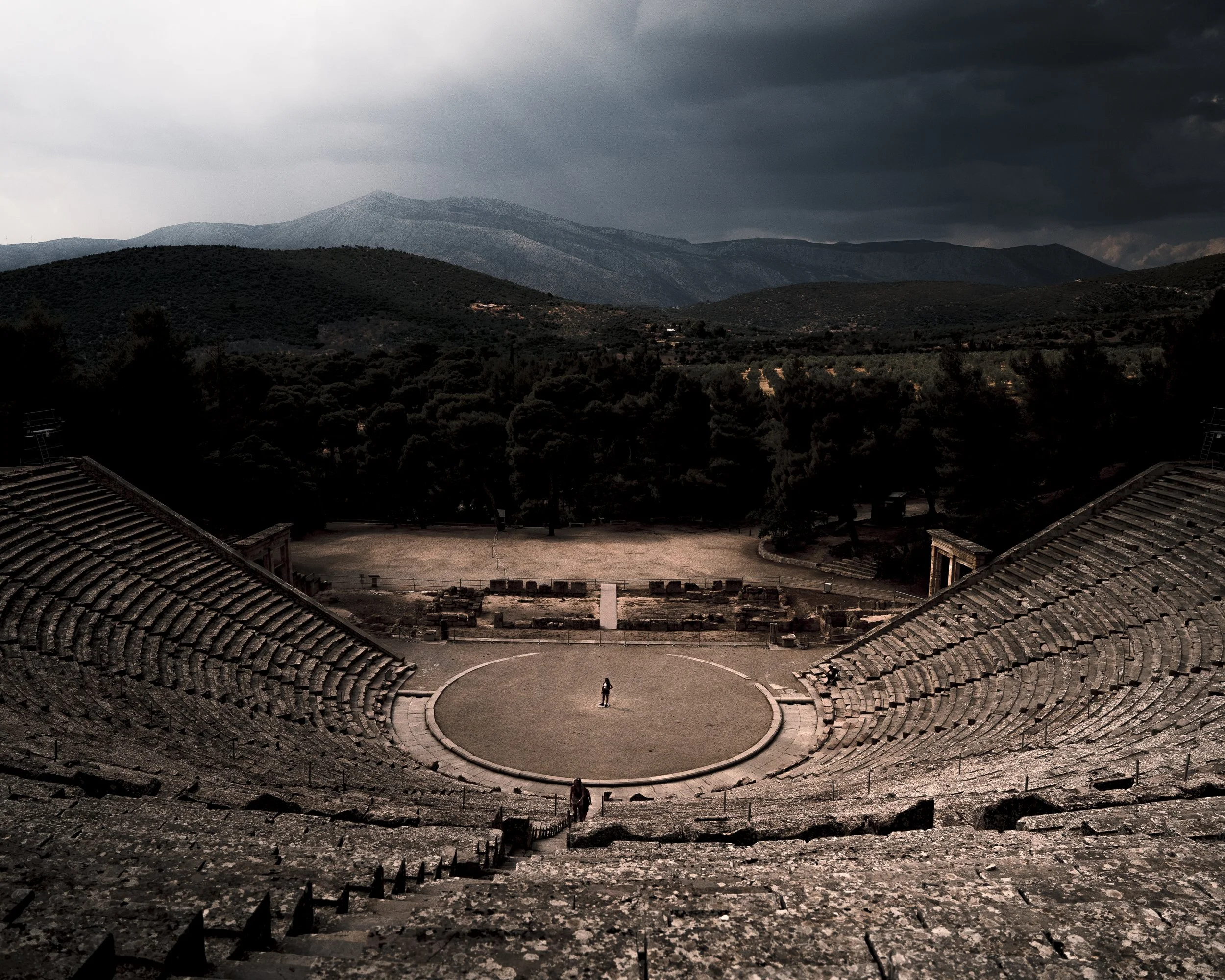 Un ancien amphithéâtre en pierre, entouré de montagnes et de végétation, sous un ciel nuageux.