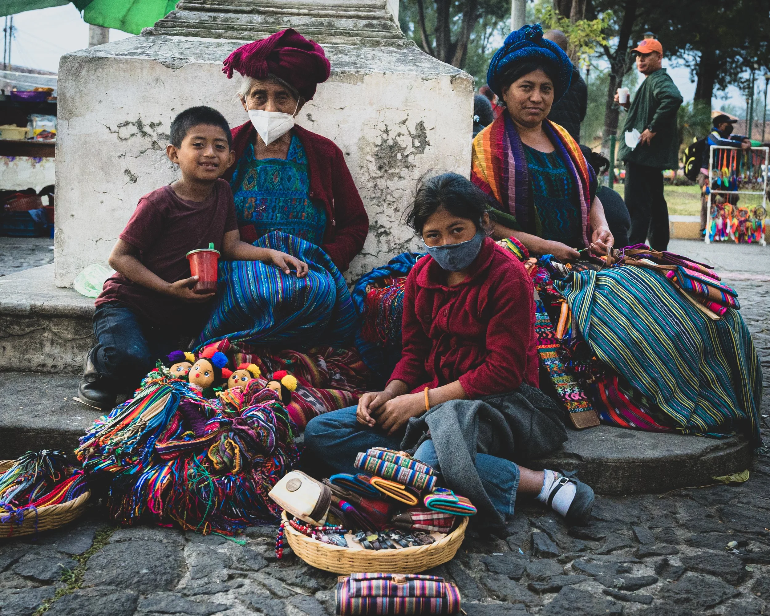 Groupe de personnes en vêtements traditionnels colorés assises sur un trottoir, vendant des articles artisanaux au marché.
