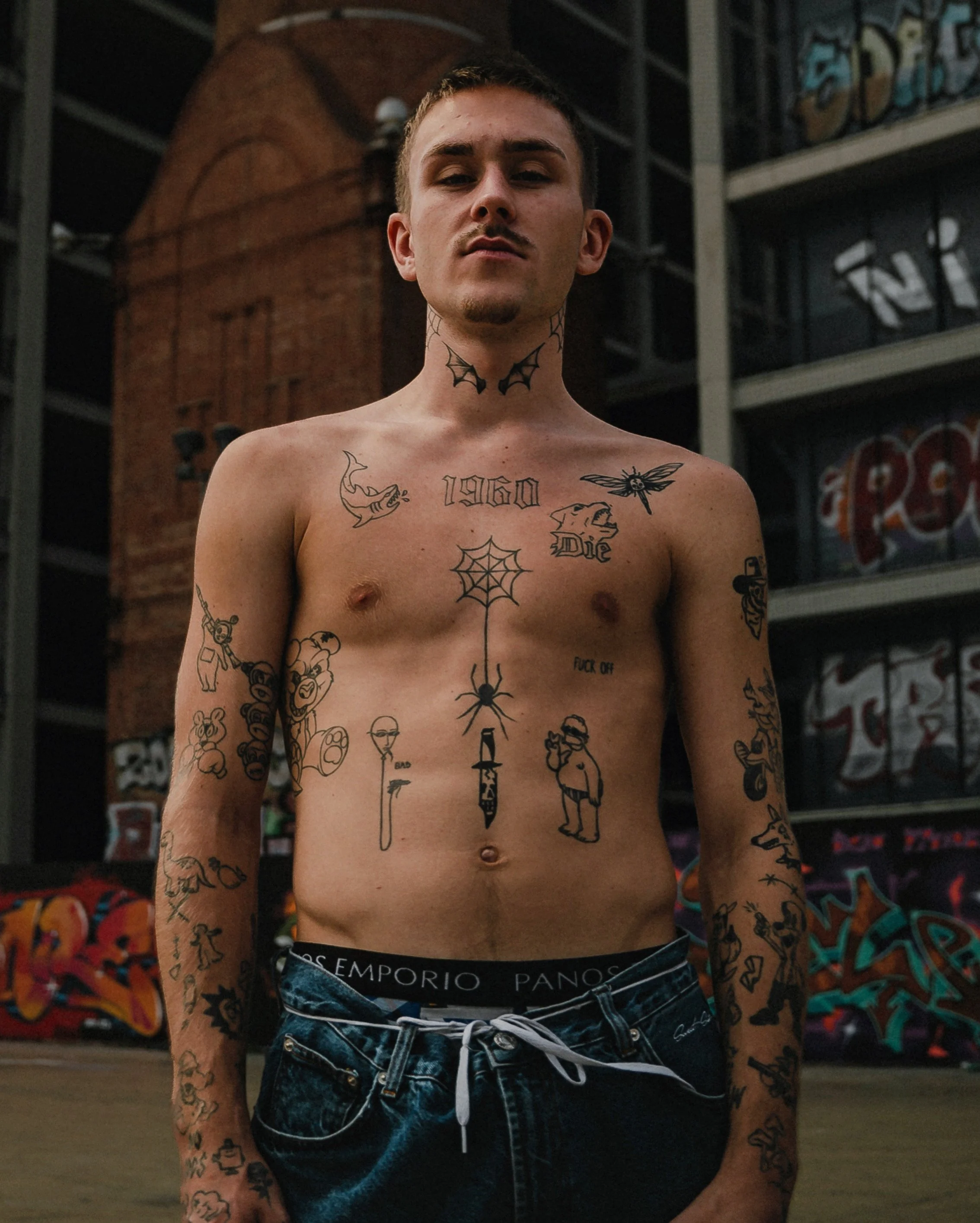 Homme torse nu avec de nombreux tatouages, debout devant un bâtiment en briques et des graffitis.