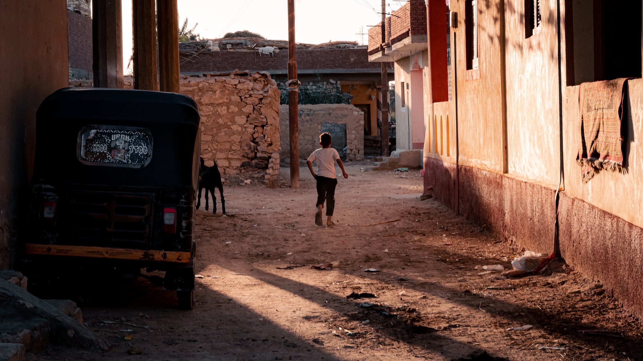 Ruelle poussiéreuse avec un enfant courant, une chèvre noire et un rickshaw garé.