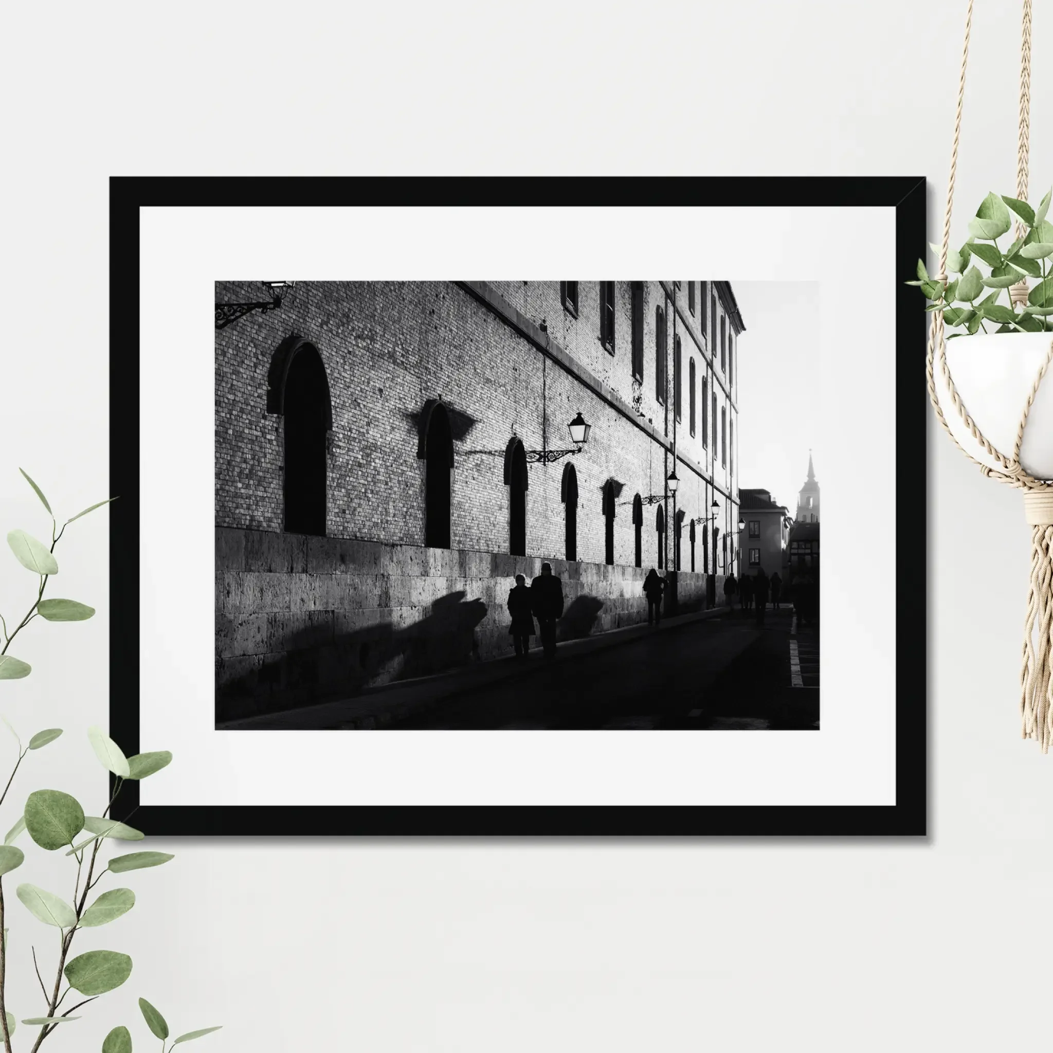 Black and white street photography print of a person walking past a textured historical wall. Features striking diagonal shadows and high-contrast lighting. Classic fine art monochrome decor.