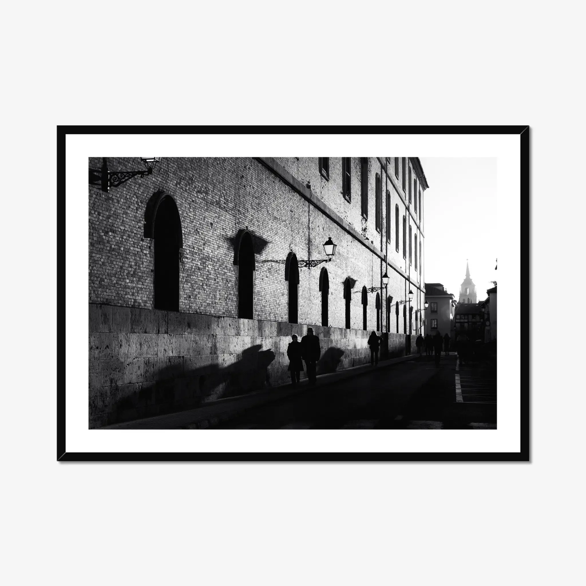 Black and white street photography print of a person walking past a textured historical wall. Features striking diagonal shadows and high-contrast lighting. Classic fine art monochrome decor.
