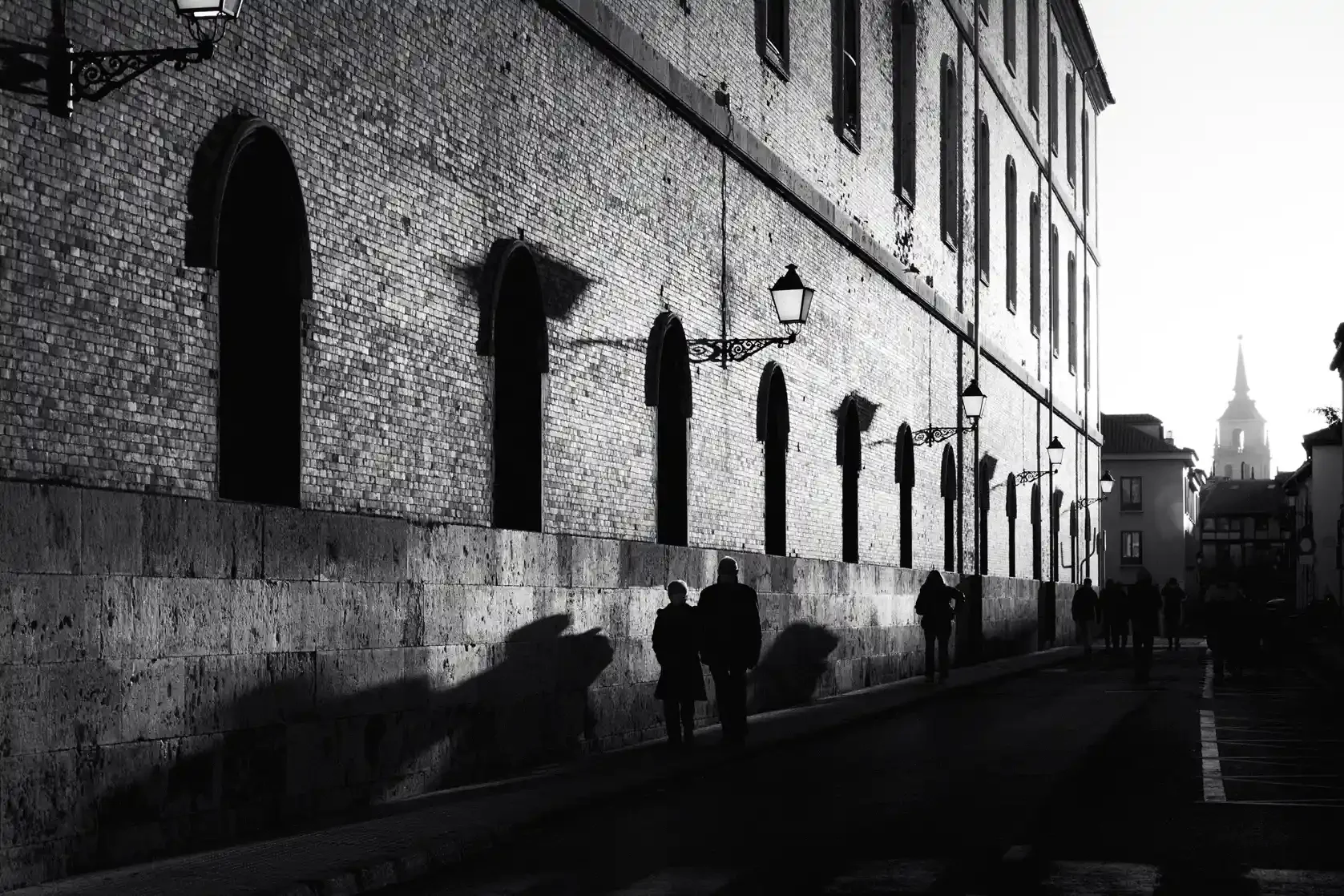 Black and white street photography print of a person walking past a textured historical wall. Features striking diagonal shadows and high-contrast lighting. Classic fine art monochrome decor.