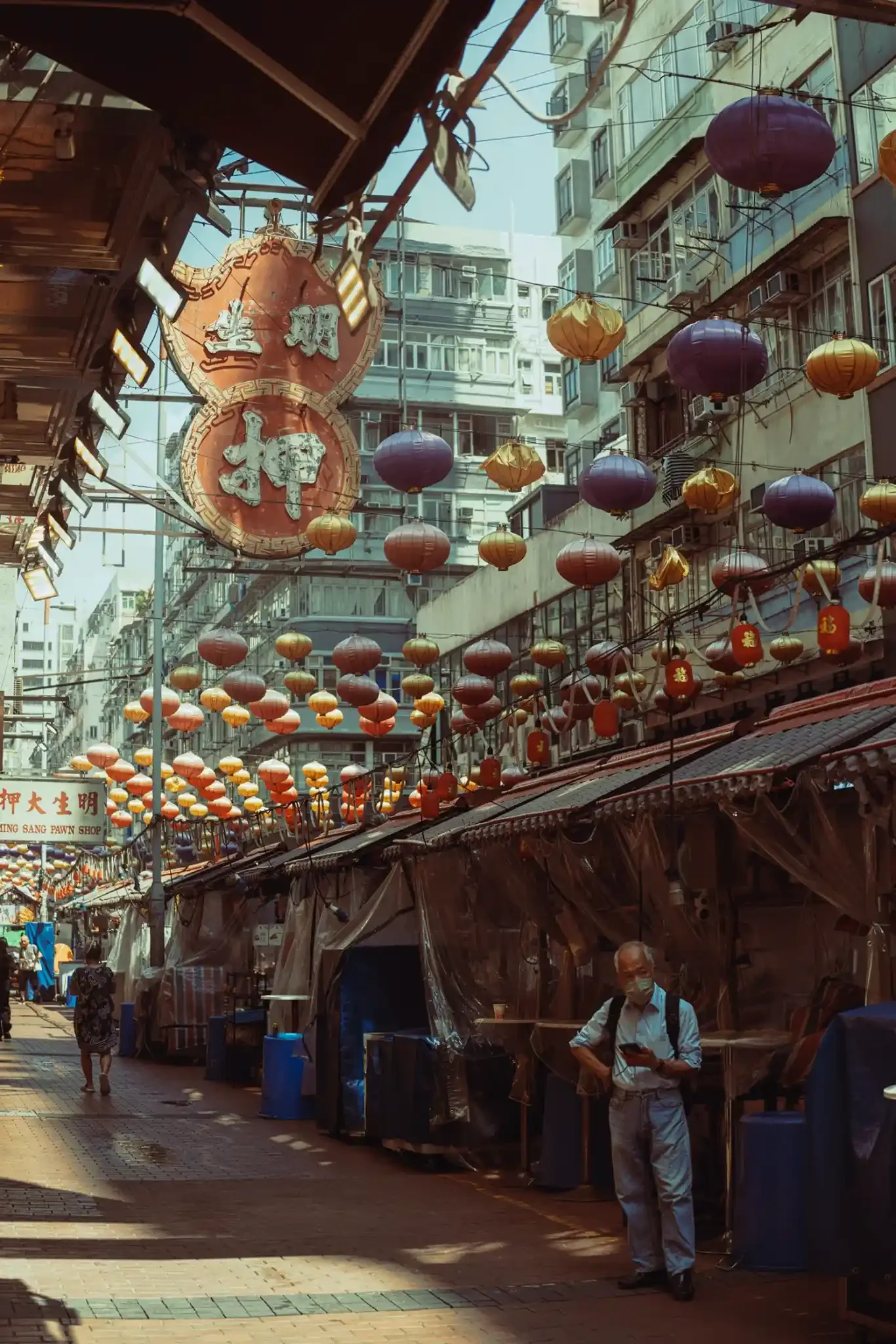 Man in a face mask checking his phone beside covered stalls on Temple Street, Hong Kong, under rows of purple, gold and red lanterns and a vintage pawn shop sign