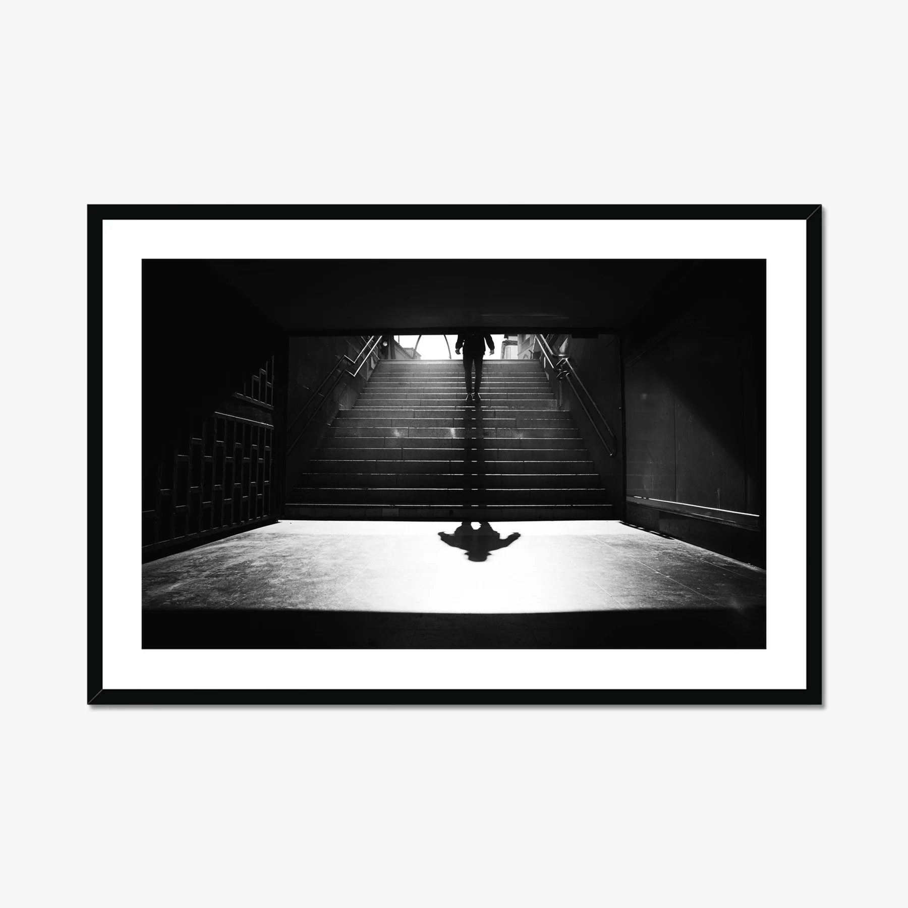 Black and white minimalist street photography of a person walking down stairs in Madrid Metro, casting a long diagonal shadow on a sunny morning.