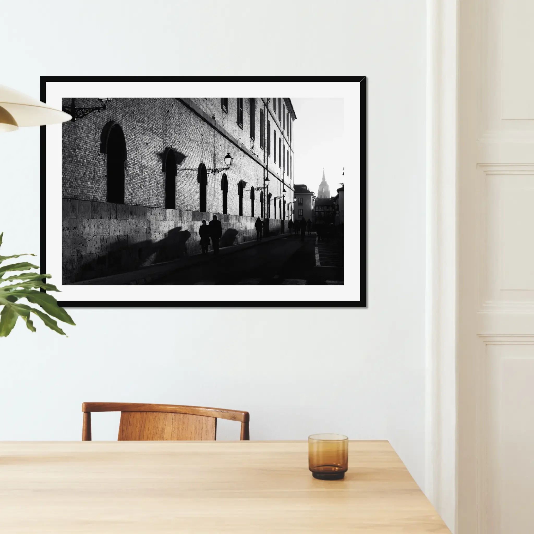 Black and white street photography print of a person walking past a textured historical wall. Features striking diagonal shadows and high-contrast lighting. Classic fine art monochrome decor.