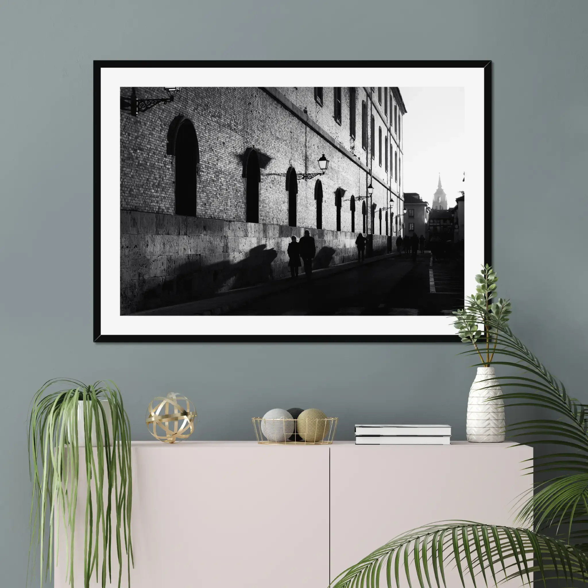 Black and white street photography print of a person walking past a textured historical wall. Features striking diagonal shadows and high-contrast lighting. Classic fine art monochrome decor.