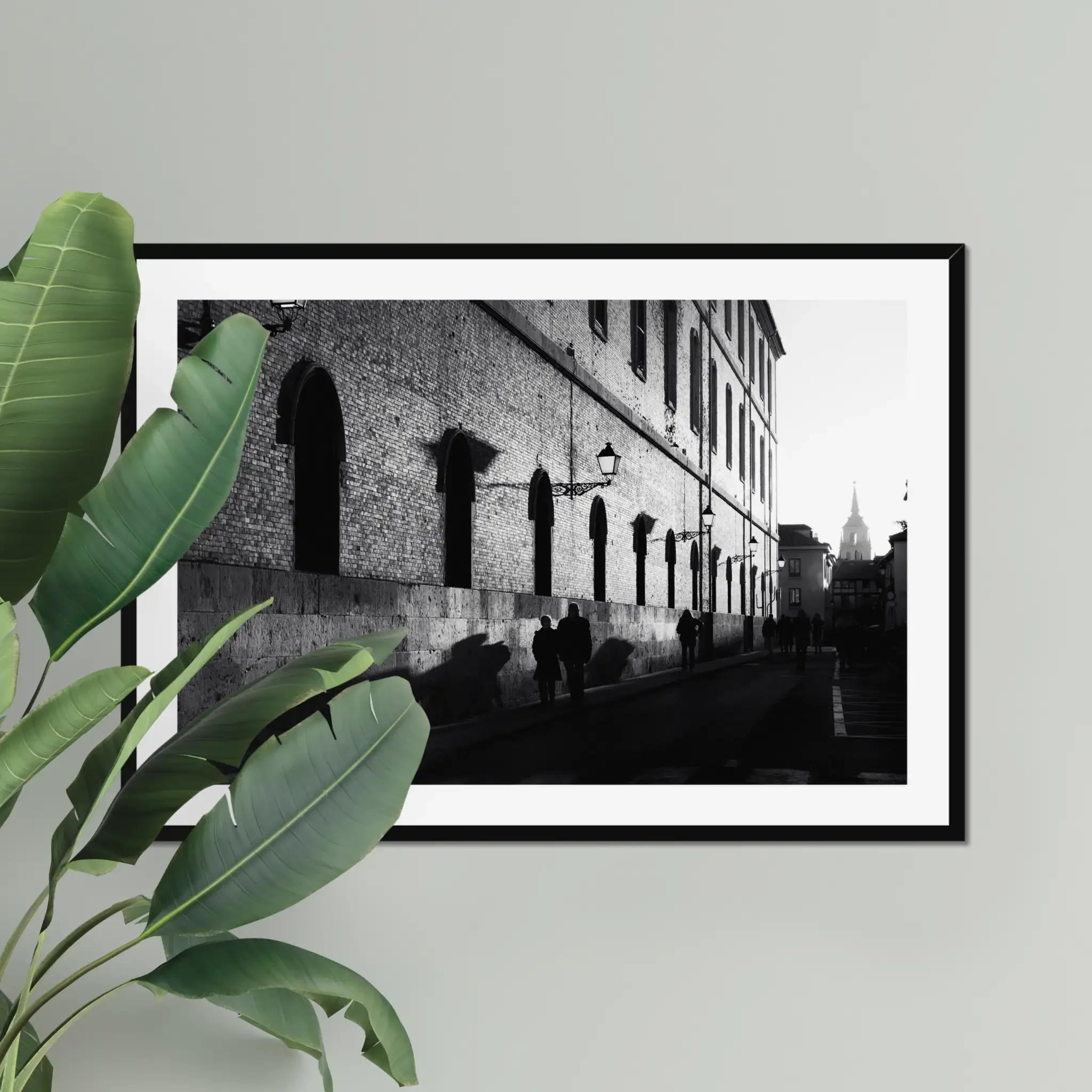 Black and white street photography print of a person walking past a textured historical wall. Features striking diagonal shadows and high-contrast lighting. Classic fine art monochrome decor.