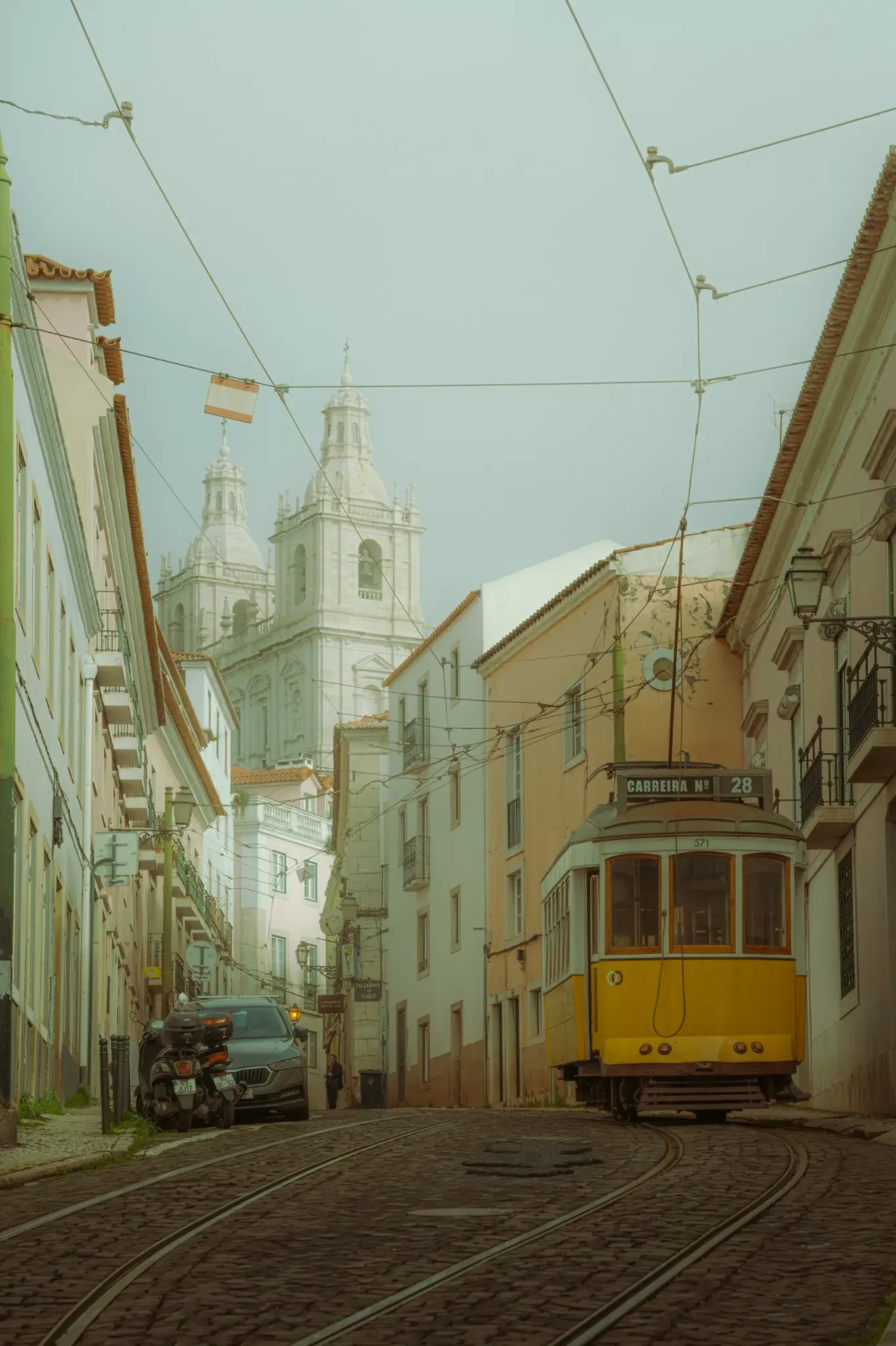 ejduranphotography Fine Art Lisbon print. Cinematic street photography of the iconic yellow Tram 28 on a winding cobblestone street with a misty church background. Moody wall art decor.