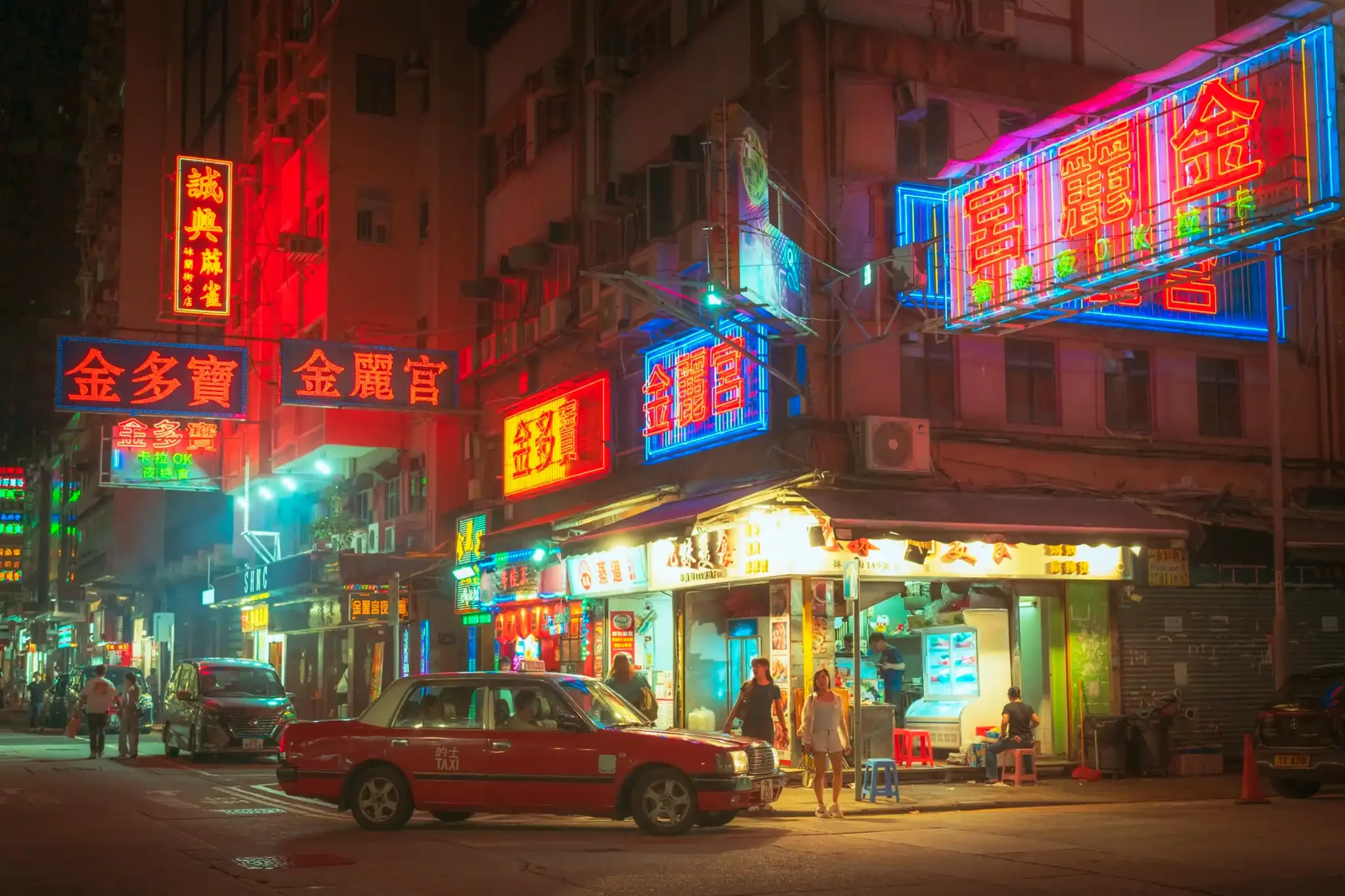 Red Hong Kong taxi parked at a night street corner beneath glowing red, blue and pink neon signs, with a street food stall and pedestrians in the background