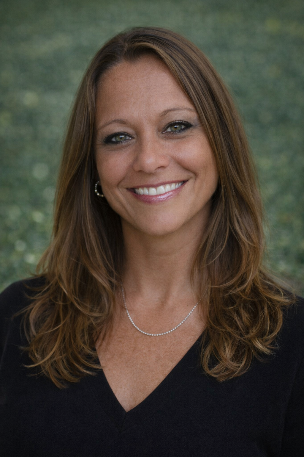 Headshot of a woman with light skin, long brown hair, gray eyes, wearing a black top, a delicate necklace, and earrings, smiling in front of a blurred green background.