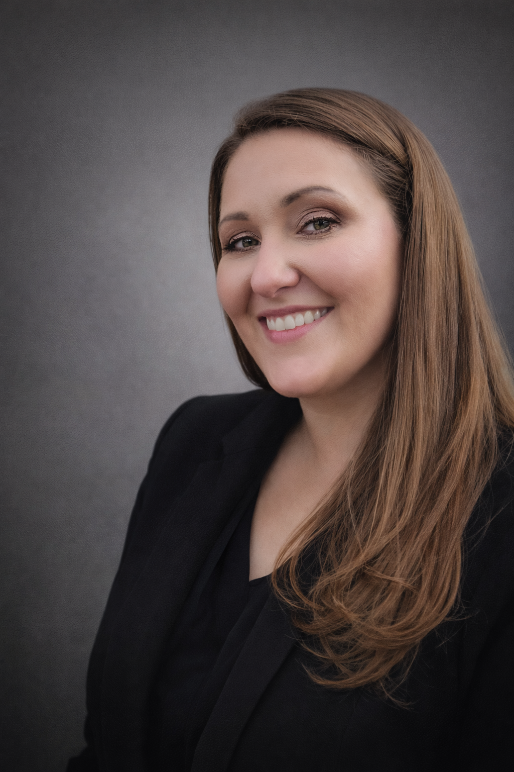 Professional portrait of a woman with long brown hair, wearing a black blazer and smiling, against a dark gray background.