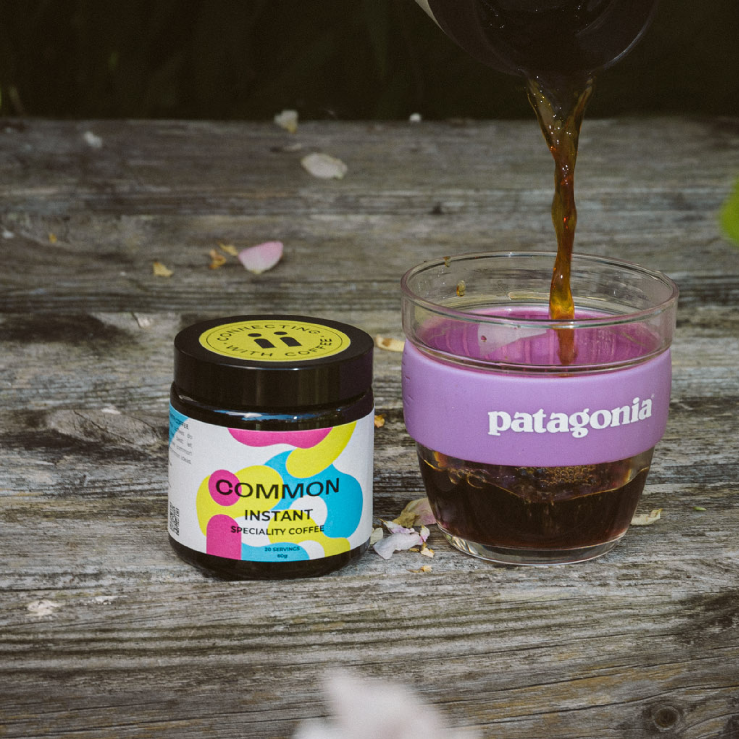 Close-up of a small jar of coffee creamer and a glass of dark iced coffee on a wooden surface, with falling flower petals and blurred background.
