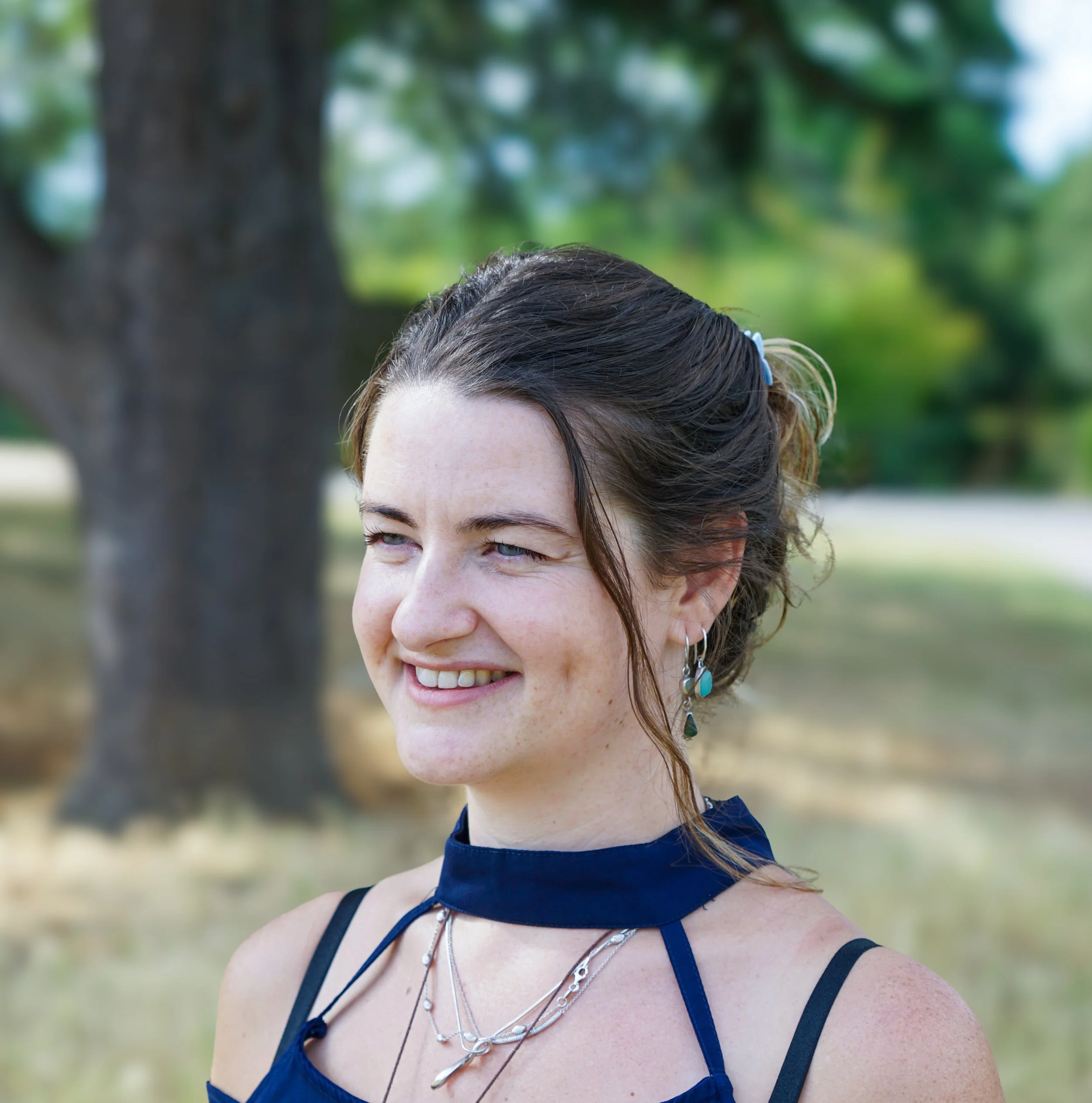 Close-up of a woman with brown hair and blue eyes, smiling, outdoors with green trees in the background. She wears layered silver necklaces, silver earrings, and a navy blue sleeveless top.