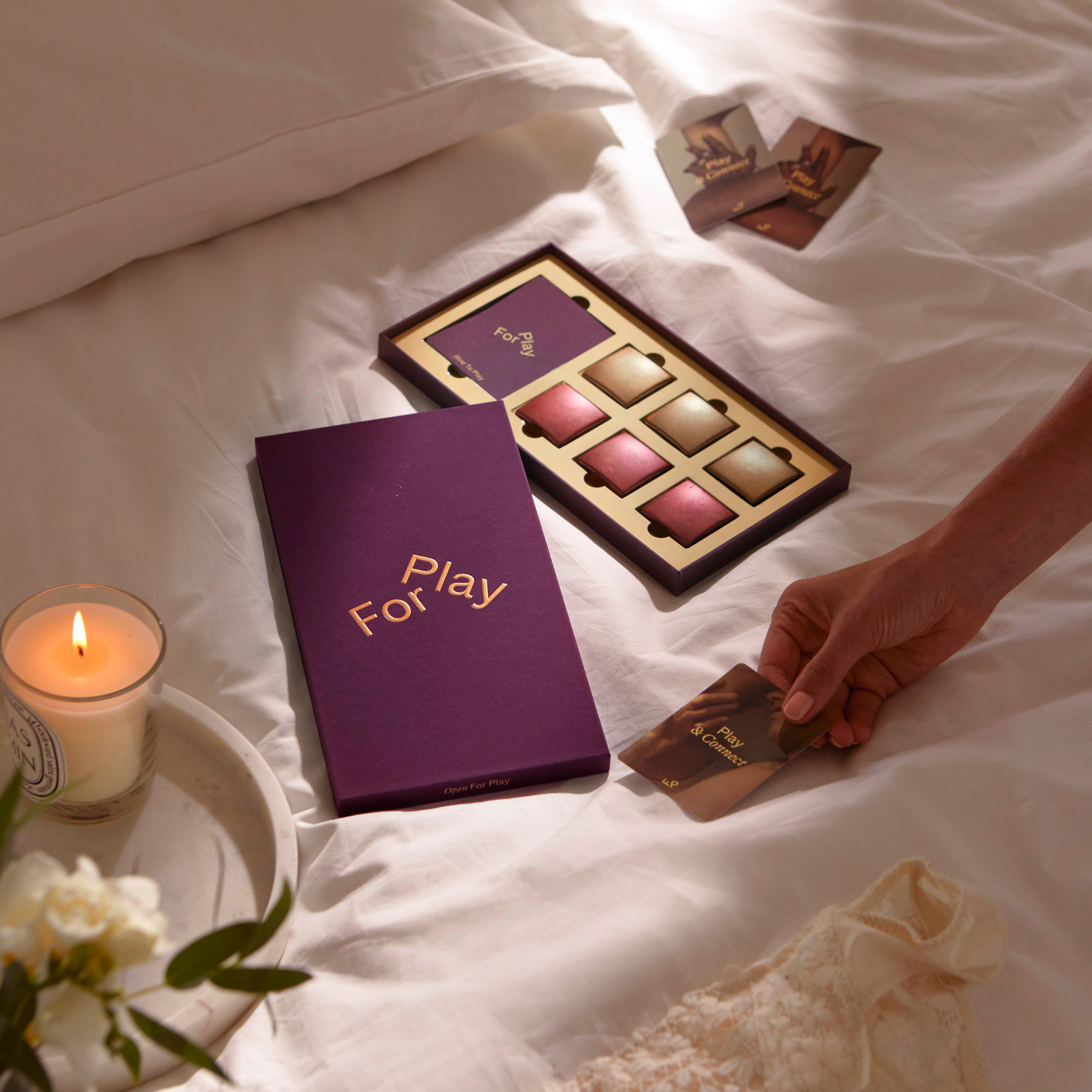 A box of makeup cosmetics with blushes, a purple box labeled 'Play For' and a woman holding a card labeled 'Play and Connect' on a white bed with a candle and flowers nearby.