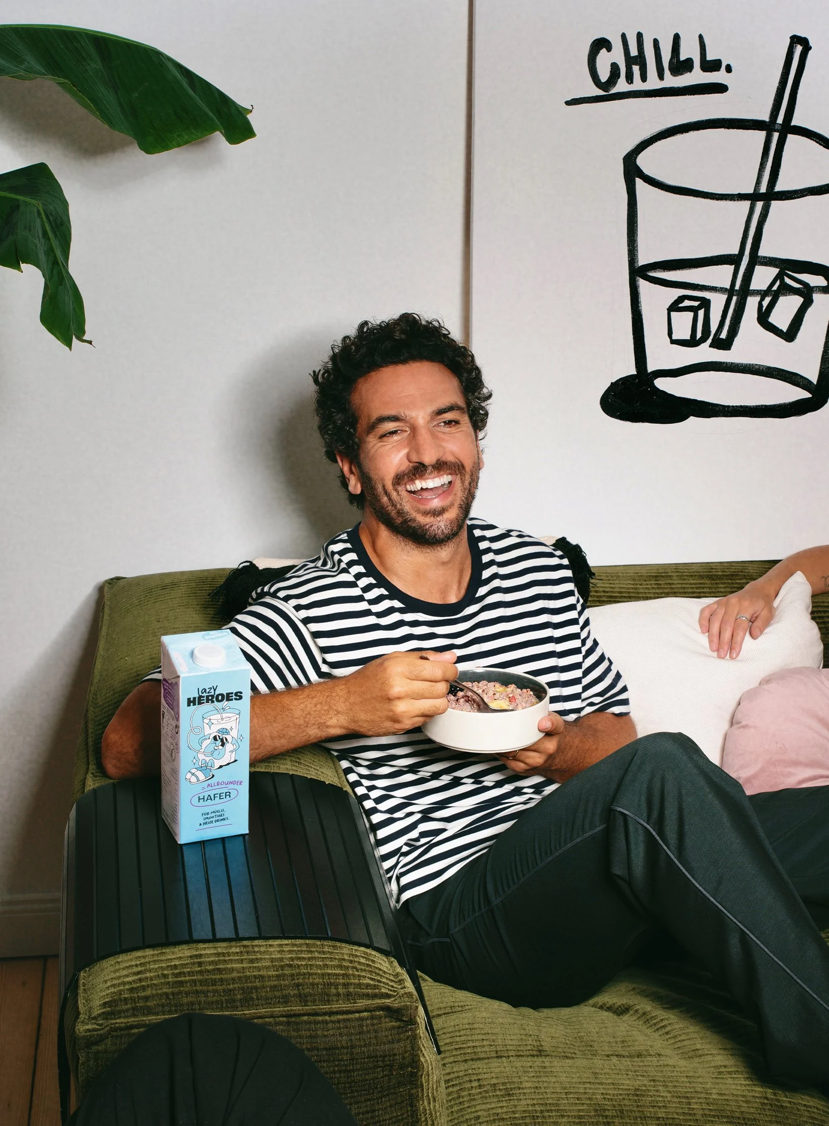 Ein lachender Mann mit dunklen, lockigen Haaren und Bart sitzt auf einem grünen Sofa, isst aus einer Schüssel, neben einer Packung Milch auf einem schwarzen Tablett, im Wohnzimmer vor einer weißen Wand mit einer schwarzen Handzeichnung eines Glases und dem Wort 'CHILL'.