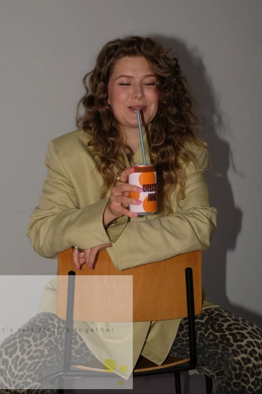 Junge Frau mit lockigem Haar trinkt aus einer Dose mit Strohhalm, sitzt auf einem Stuhl vor grauem Hintergrund, trägt einen beigen Blazer und leopardene Leggings.