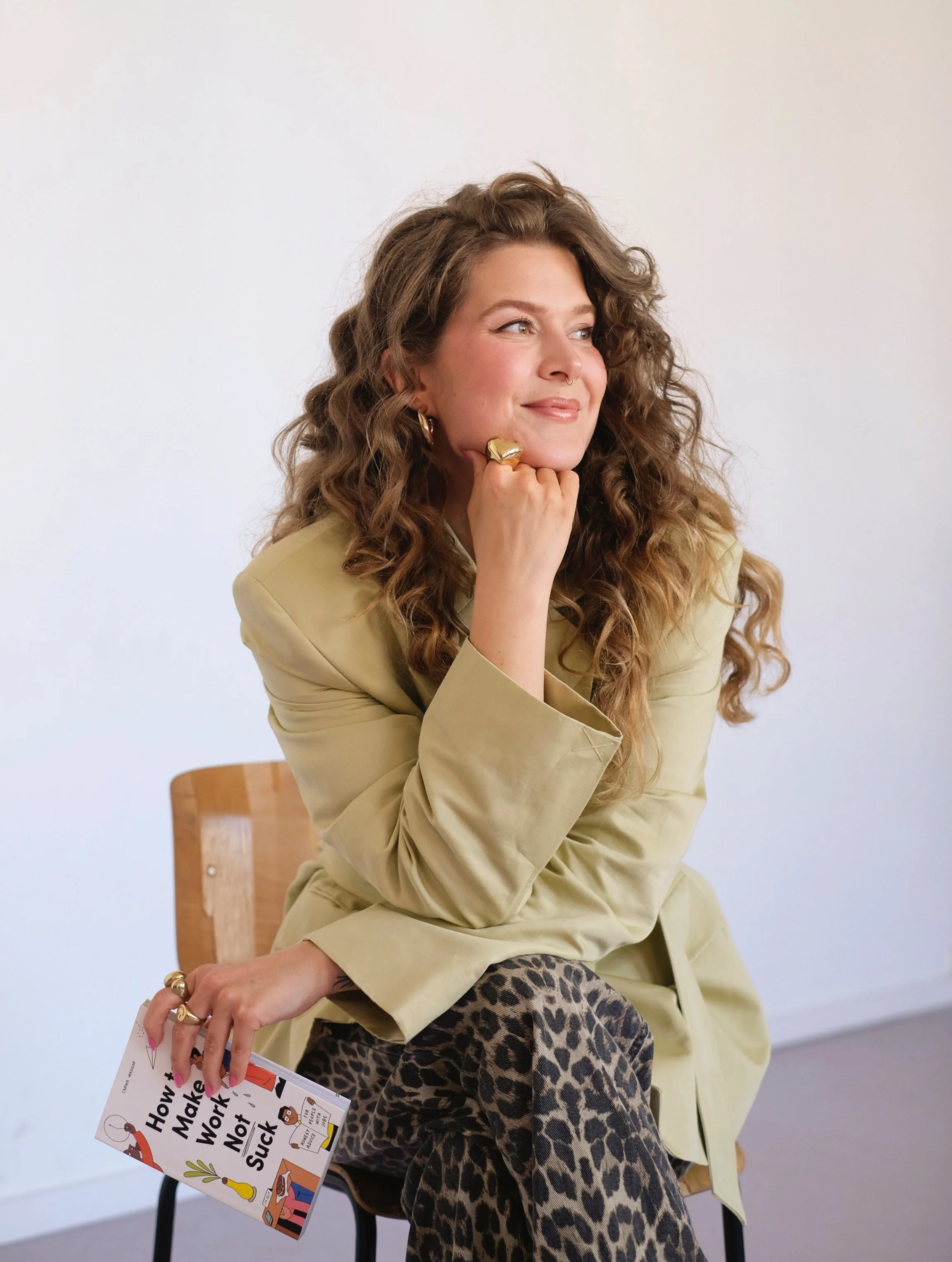 Eine junge Frau mit langen, lockigen Haaren sitzt auf einem Stuhl und lächelt nach rechts. Sie trägt eine beige Jacke, Leopardenmuster-Hosen und mehrere goldene Ringe. In ihrer rechten Hand hält sie ein Buch mit dem Titel 'How to Make Work Not Suck'.