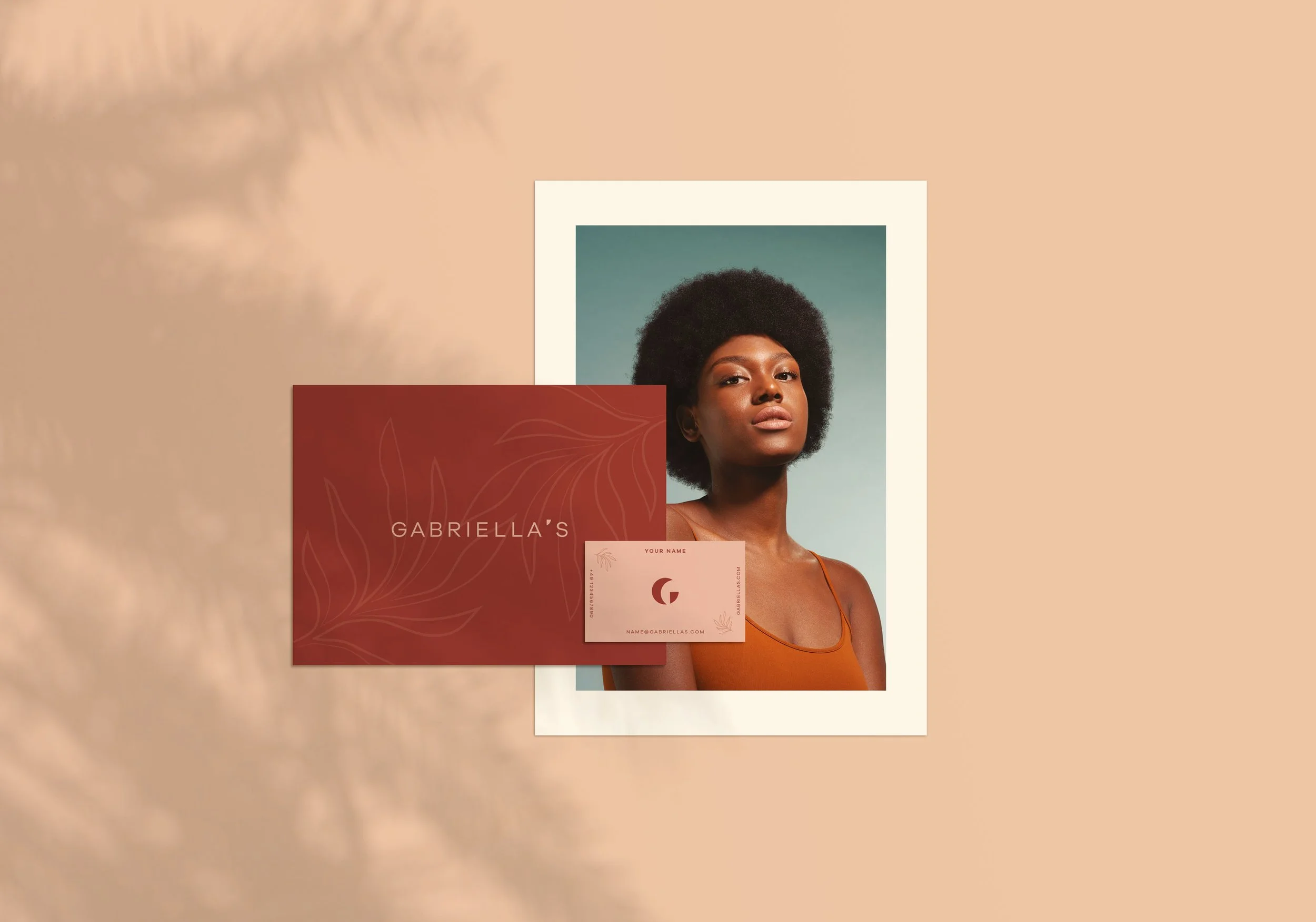 Eine Fotografie einer schwarzen Frau mit Afro-Haar, die ein braunes Oberteil trägt, vor einem grauen Hintergrund. Im Vordergrund liegt eine rote Visitenkarte mit Blattmuster, die den Text "GABRIELLA'S" enthält, sowie eine kleine Karte mit Platz für den Namen, E-Mail und Webseite.