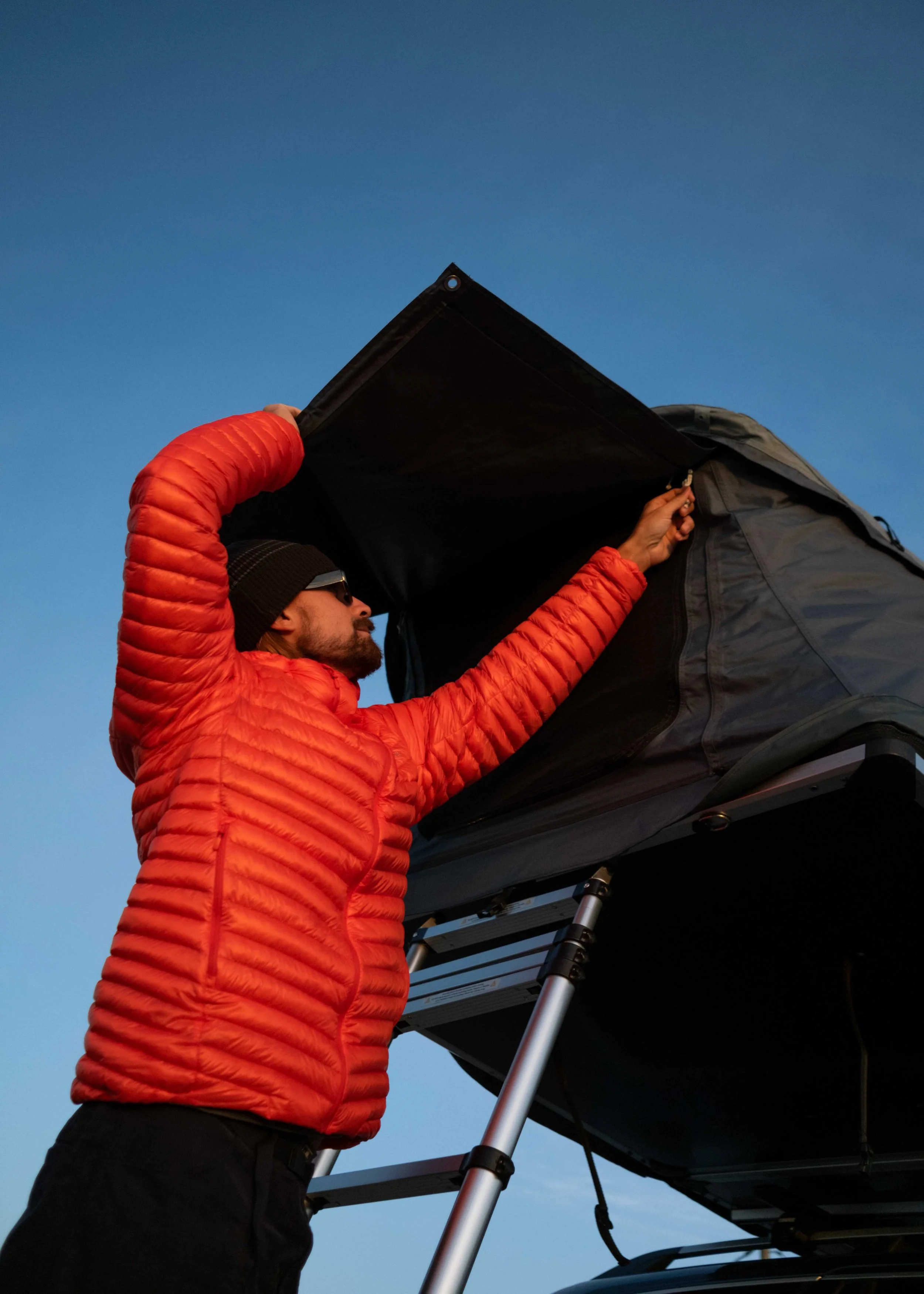 A man in a red jacket, black beanie, and sunglasses is adjusting equipment on top of a black rooftop tent mounted on a vehicle during sunset or sunrise.