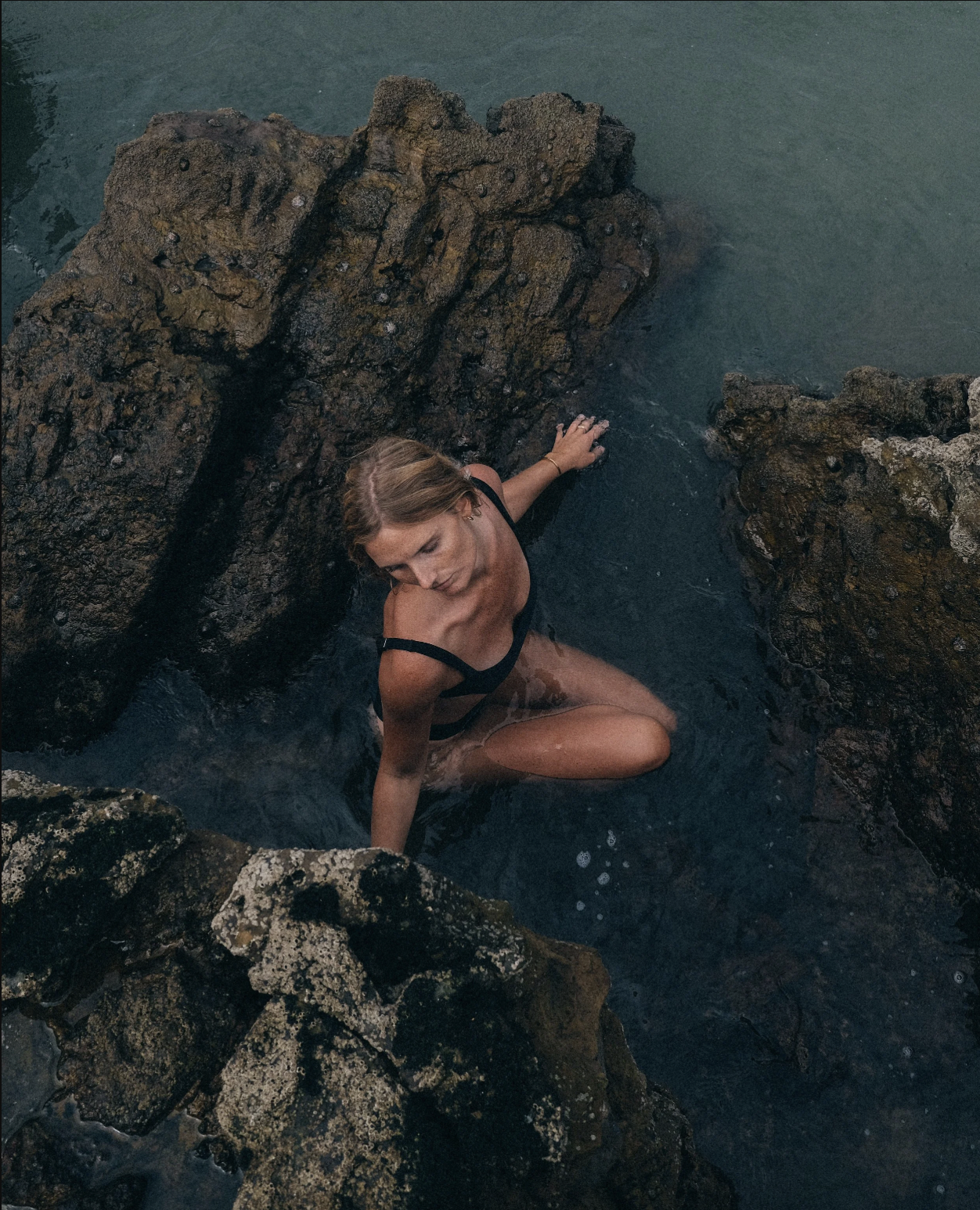Woman in a black swimsuit kneeling in a rocky coastal area with water around her, looking down.