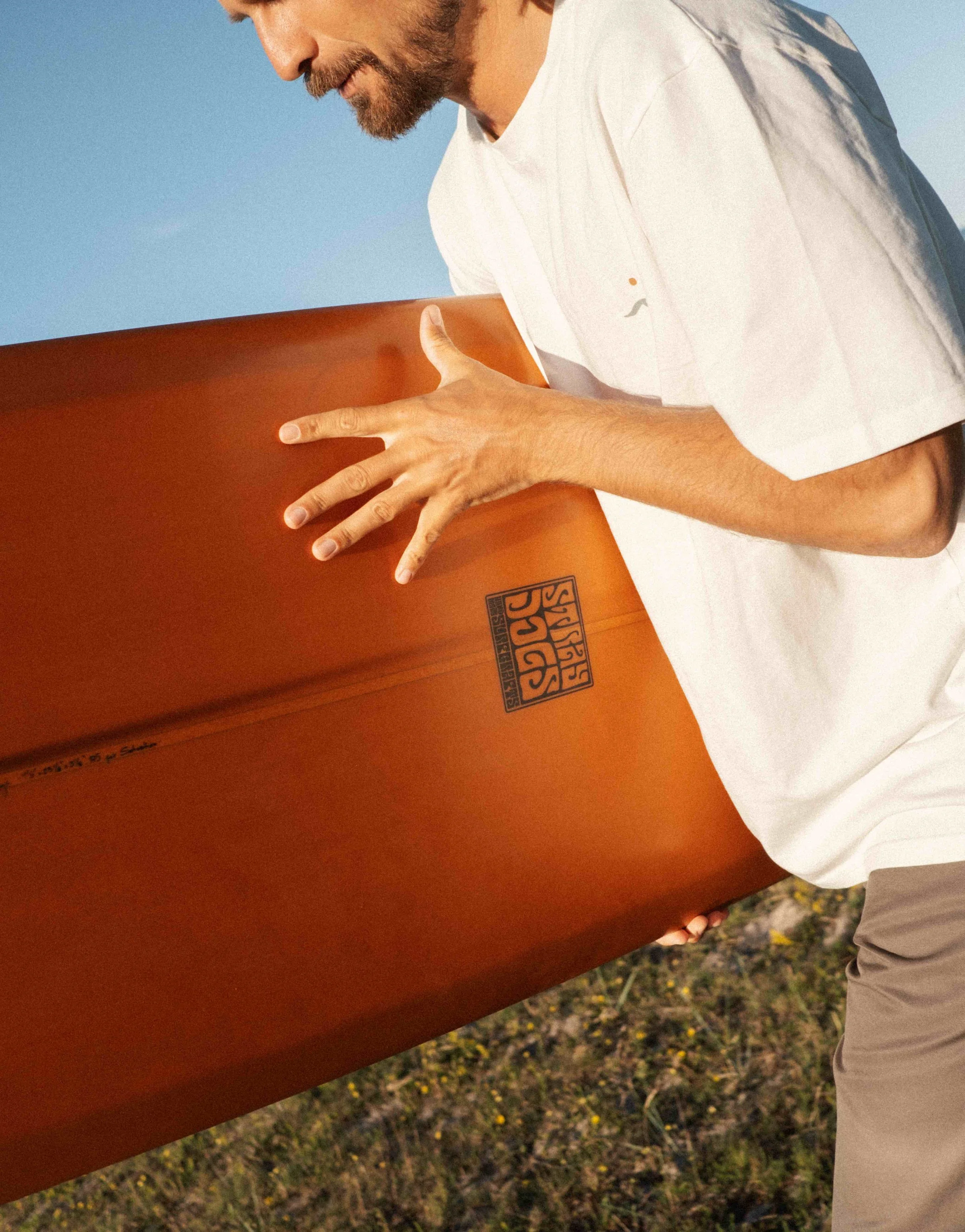 A man holding a surfboard on a grassy and rocky land area, with a clear blue sky in the background.