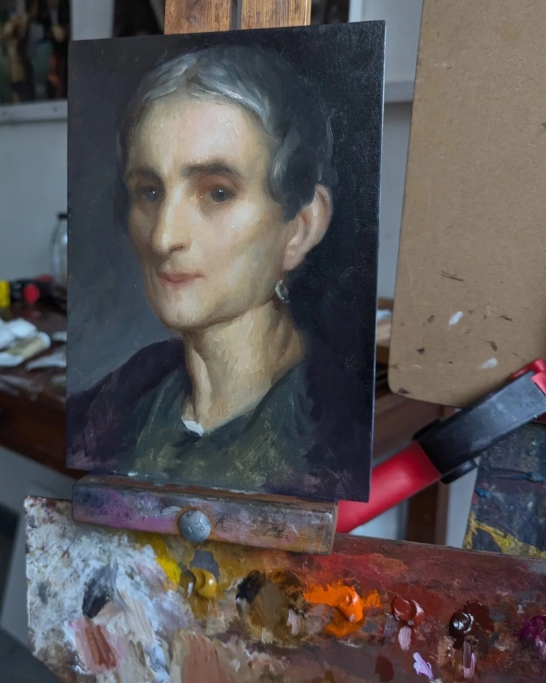 A portrait painting of a woman with short, dark hair, wearing earrings, and dressed in dark clothing, on an easel in an artist's studio. The palette below shows mixed paint colors.
